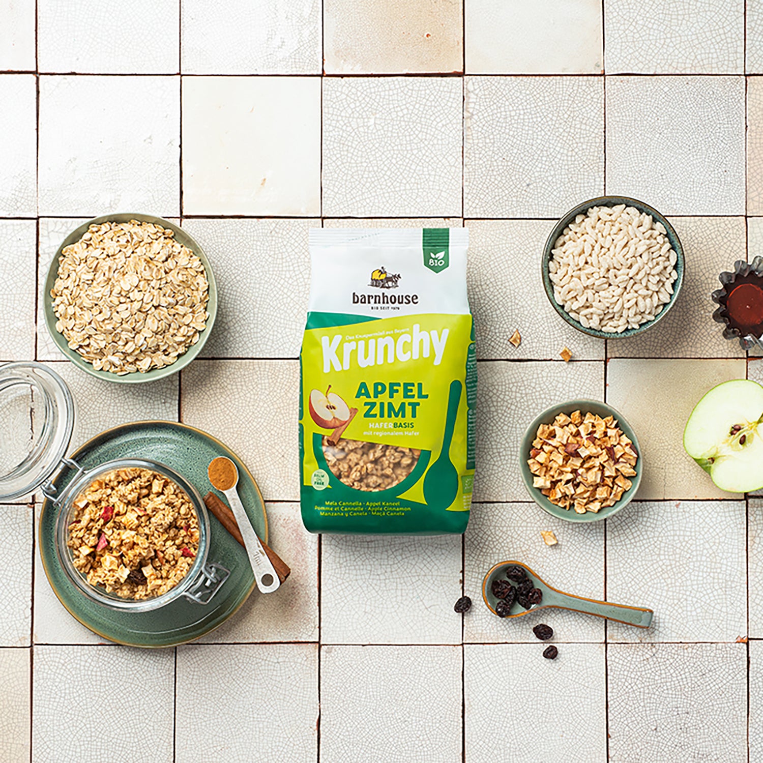 Beutel Krunchy Apfel-Zimt von Bio-Knuspermüsli-Hersteller Barnhouse auf gemusterten Fließen. Links steht ein grünen Teller mit durchsichtigem Gefäß & Krunchy Apfel-Zimt sowie eine Zimtstange & ein Löffel mit Zimt. Zudem stehen dort noch 3 grünliche Schalen mit den Zutaten Haferflocken, Apfelstückchen und Knusperreis. Vereinzelte Apfelstückchen sind auf dem Tisch verteilt & unten rechts liegen noch ein paar Rosinen auf dem Tisch sowie in einem grün-braunen Löffel. Rechts eine angeschnittene Apfelhälfte.