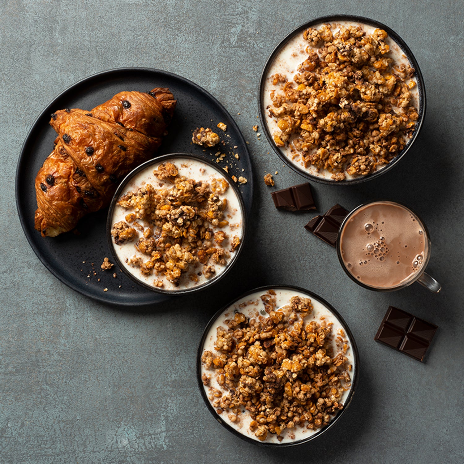 Blick von oben auf einen gräulichen Tisch mit einem schwarzen Teller, auf dem ein Schoko-Croissant sowie eine Schüssel mit Milch und Krunchy Chocolate Chunks von Bio-Knuspermüsli-Hersteller Barnhouse zu sehen sind. Daneben stehen 2 Schüsseln, die jeweils mit diesem Müsli und Milch befüllt sind. Außerdem befinden sich noch eine Tasse heiße Schokolade und dunkle Schoko-Stückchen auf dem Tisch.