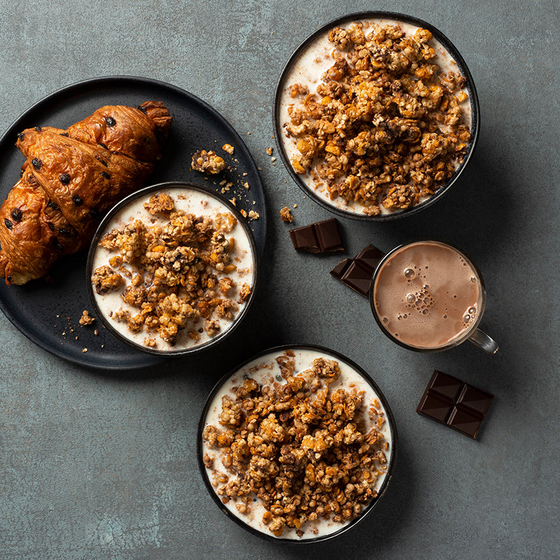 Blick von oben auf einen gräulichen Tisch mit einem schwarzen Teller, auf dem ein Schoko-Croissant sowie eine Schüssel mit Milch und Krunchy Chocolate Chunks von Bio-Knuspermüsli-Hersteller Barnhouse zu sehen sind. Daneben stehen 2 Schüsseln, die jeweils mit diesem Müsli und Milch befüllt sind. Außerdem befinden sich noch eine Tasse heiße Schokolade und dunkle Schoko-Stückchen auf dem Tisch.
