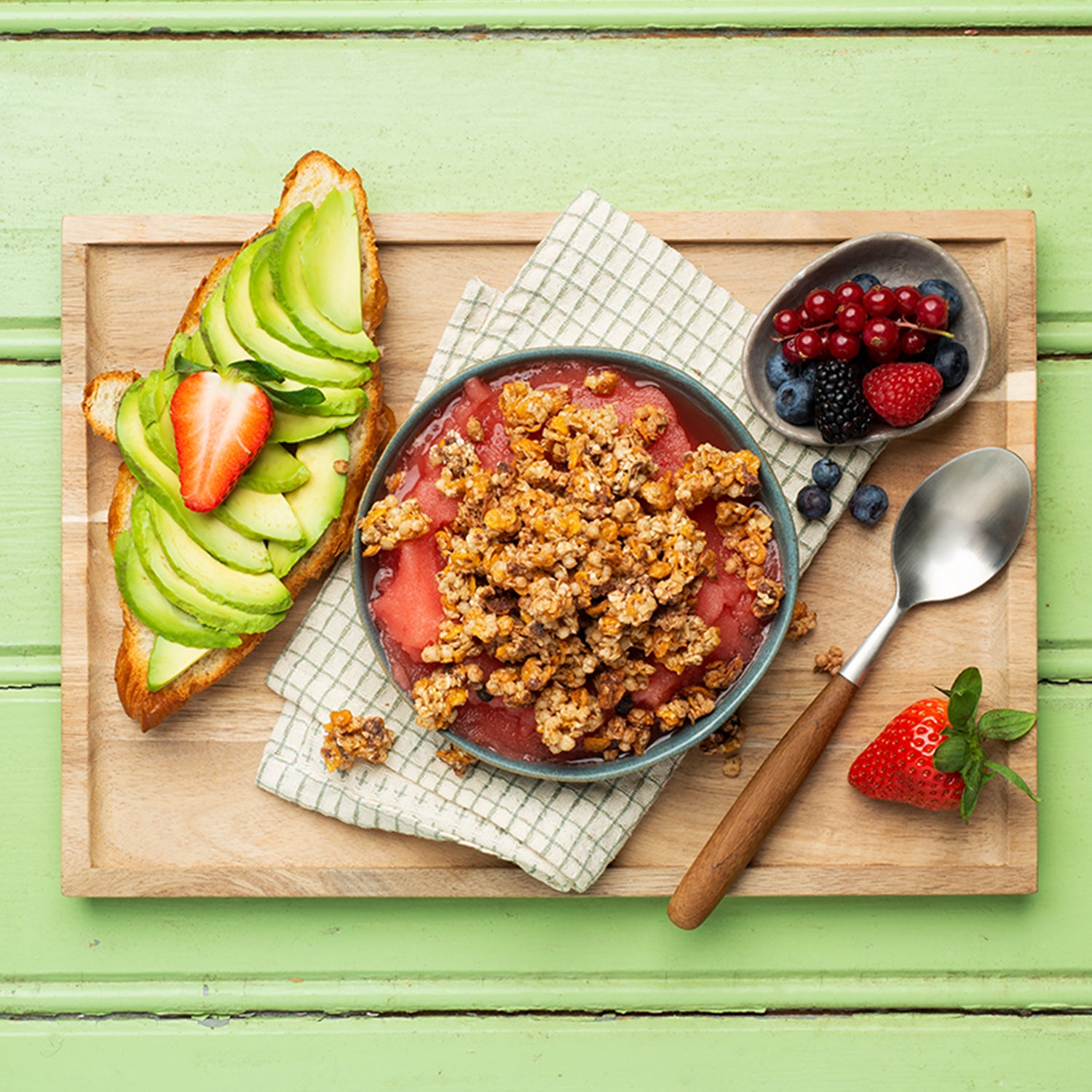 Ein grüner Holztisch, auf dem eine braune Holzplatte liegt. Darauf sind ein Weißbrot mit Avocado und Erdbeere sowie eine Schüssel mit Beeren und eine weitere Erdbeere zu sehen. Außerdem liegen noch ein Löffel, Cluster dieses Müslis und eine karierte Serviette auf dem Brett, auf der wiederum eine Schüssel mit Erdbeere-Mousse und diesem Krunchy steht.