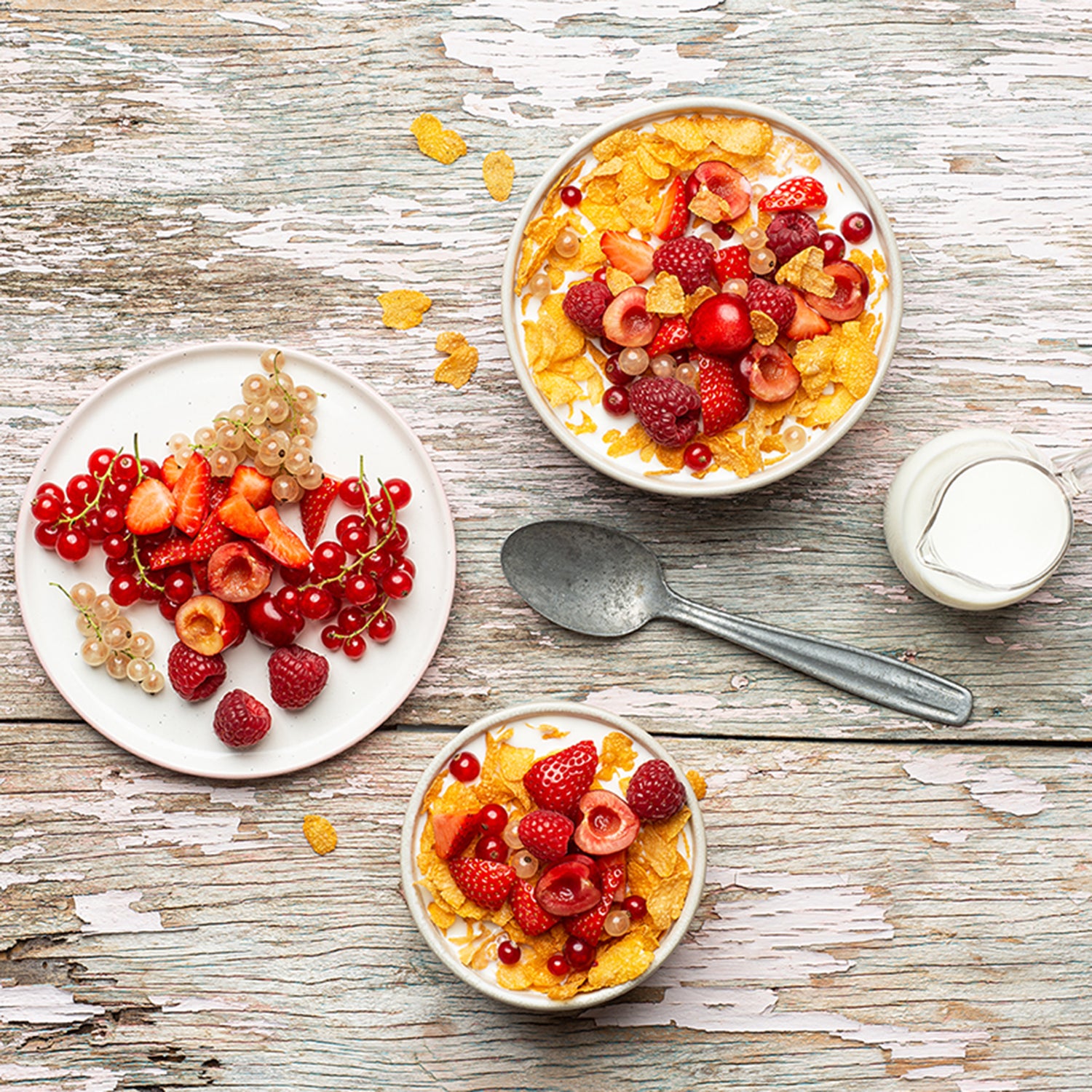 Holztisch, der weiß gestrichen ist und schon an einigen Stellen abblättert. Darauf befinden sich ein Teller mit einer Beeren-Auswahl sowie 2 Schüsseln, die jeweils mit Cornflakes Original von Bio-Knuspermüsli-Hersteller Barnhouse befüllt sind. Auch darin befindet sich eine bunte Auswahl an Beeren. Zudem liegen noch ein grauer Löffel sowie eine kleine Kanne mit Milch auf dem Tisch.