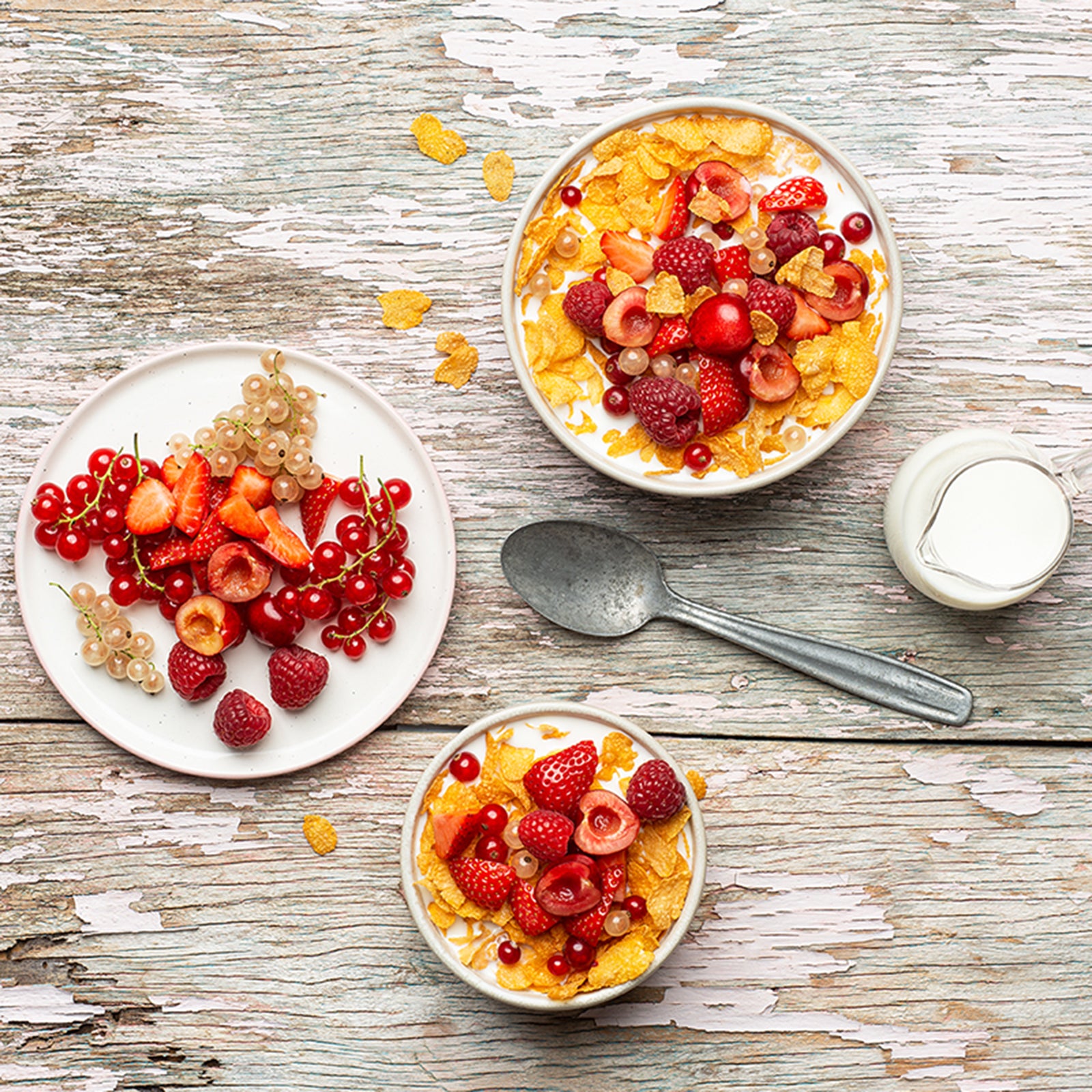 Holztisch, der weiß gestrichen ist und schon an einigen Stellen abblättert. Darauf befinden sich ein Teller mit einer Beeren-Auswahl sowie 2 Schüsseln, die jeweils mit Cornflakes Original von Bio-Knuspermüsli-Hersteller Barnhouse befüllt sind. Auch darin befindet sich eine bunte Auswahl an Beeren. Zudem liegen noch ein grauer Löffel sowie eine kleine Kanne mit Milch auf dem Tisch.