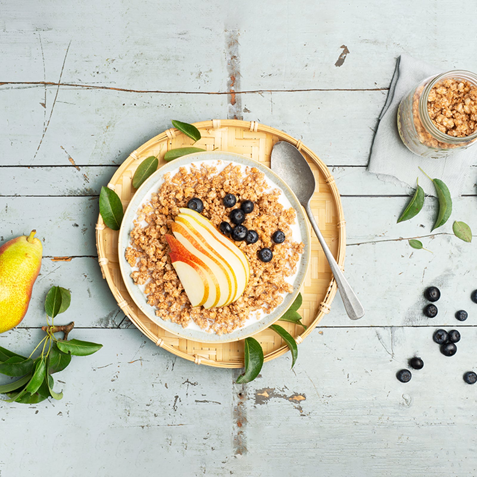 Ein etwas abgenutzter, gräulicher Holztisch mit einem geflochtenen Unterteller. Darauf liegen ein paar Blätter sowie eine weiße Schüssel, die mit Krunchy Dinkel von Bio-Knuspermüsli Hersteller Barnhouse befüllt ist. Als Topping liegen ein paar Apfelscheiben und Heidelbeeren drauf. Auf dem Tisch liegen noch eine Birne, weitere Blätter und paar Heidelbeeren sowie ein Glas mit besagtem Müsli.