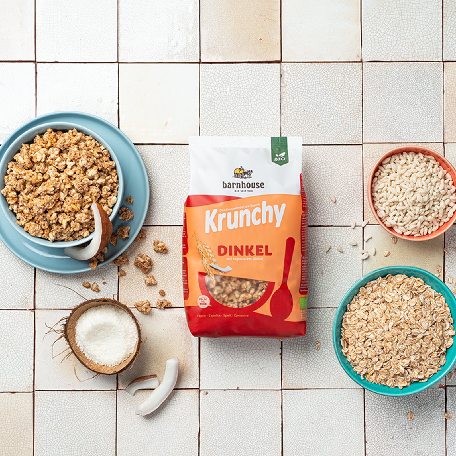 Beutel Krunchy Dinkel von Bio-Knuspermüsli-Hersteller Barnhouse auf gemusterten Fließen. Links steht ein blauer Teller mit blauer Schüssel, befüllt mit diesem Müsli. Kokosnuss-Stücke und Müsli-Cluster liegen auf dem Unterteller sowie auf den Fließen verteilt. Vorne links steht eine halbe Kokosnuss mit Meersalz befüllt. Auf der rechten Seite steht eine rote Schüssel mit Knusperreis und eine blaue Schüssel mit Haferflocken. Zwischen den beiden Schalen liegen noch ein paar weitere Haferflocken & Knusperreis.