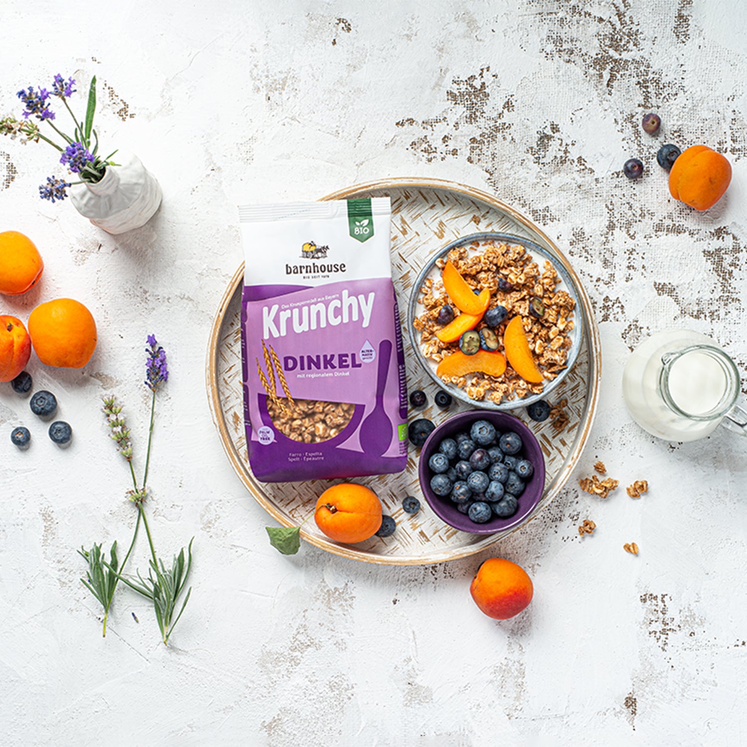 Etwas abgenutzter, weißer Holztisch mit geflochtenem Unterteller. Darauf liegt ein violetter Beutel Krunchy Dinkel alternativ gesüßt von Bio-Knuspermüsli Hersteller Barnhouse. Daneben eine blaue Schüssel mit Milch, dem besagten Müsli, Aprikosen-Stücken und Heidelbeeren. Darunter steht eine violette Schale mit einigen Heidelbeeren. Auf dem Tisch sowie dem Unterteller sind noch ein paar Müsli-Cluster, Aprikosen und Heidelbeeren verteilt. Außerdem auf dem Tisch: Glaskaraffe mit Milch + Blumen (in einer Vase).
