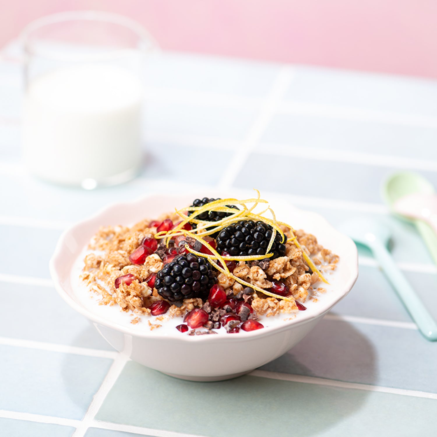 Der Fokus liegt auf einer weißen Schale, die mit Milch und Krunchy Dinkel alternativ gesüßt von Bio-Knuspermüsli-Hersteller Barnhouse gefüllt ist. Die Bowl ist verfeinert mit Johannisbeeren, Brombeeren und Zitronenzesten. Im Hintergrund sind noch etwas unscharf ein Glas Milch sowie ein grüner und ein blauer Löffel zu sehen. Die Tischdecke hat das Design einer hellblauen, gemauerten Wand. Die Wand hinter dem Tisch ist rosa.