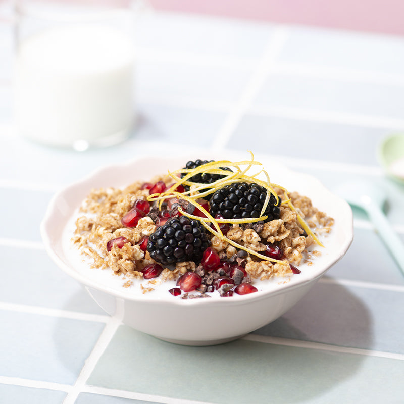Der Fokus liegt auf einer weißen Schale, die mit Milch und Krunchy Dinkel alternativ gesüßt von Bio-Knuspermüsli-Hersteller Barnhouse gefüllt ist. Die Bowl ist verfeinert mit Johannisbeeren, Brombeeren und Zitronenzesten. Im Hintergrund sind noch etwas unscharf ein Glas Milch sowie ein grüner und ein blauer Löffel zu sehen. Die Tischdecke hat das Design einer hellblauen, gemauerten Wand. Die Wand hinter dem Tisch ist rosa.