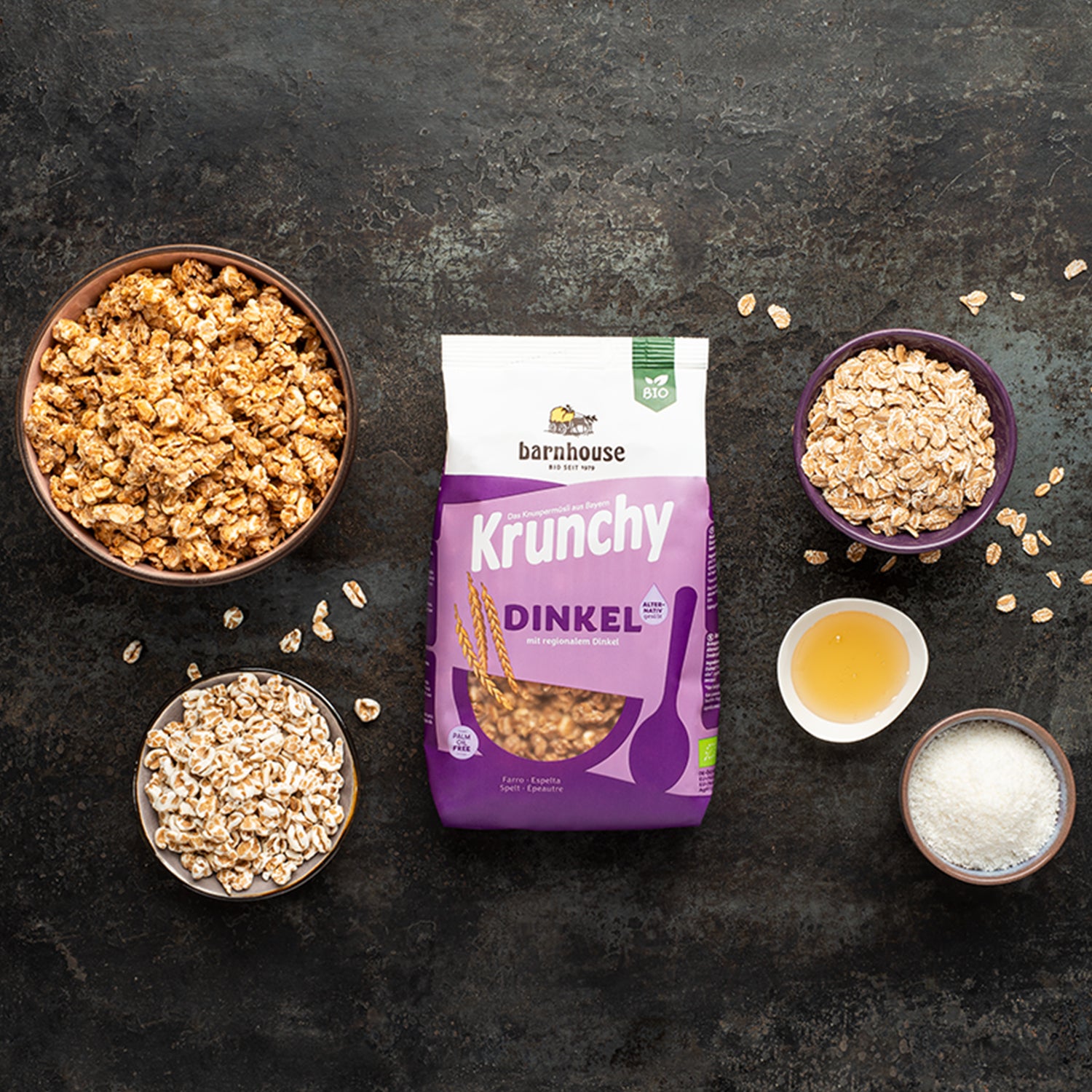 Beutel Krunchy Dinkel alternativ gesüßt von Bio-Knuspermüsli-Hersteller Barnhouse auf schwarzer Marmorplatte. Außenrum verschiedenfarbige Schüsseln mit Müsli-Clustern sowie den Zutaten Dinkelflocken, gepuffter Dinkel, Kokosraspeln und Reissirup. Auf dem Tisch sind noch ein paar Dinkelflocken sowie gepuffter Dinkel verteilt.