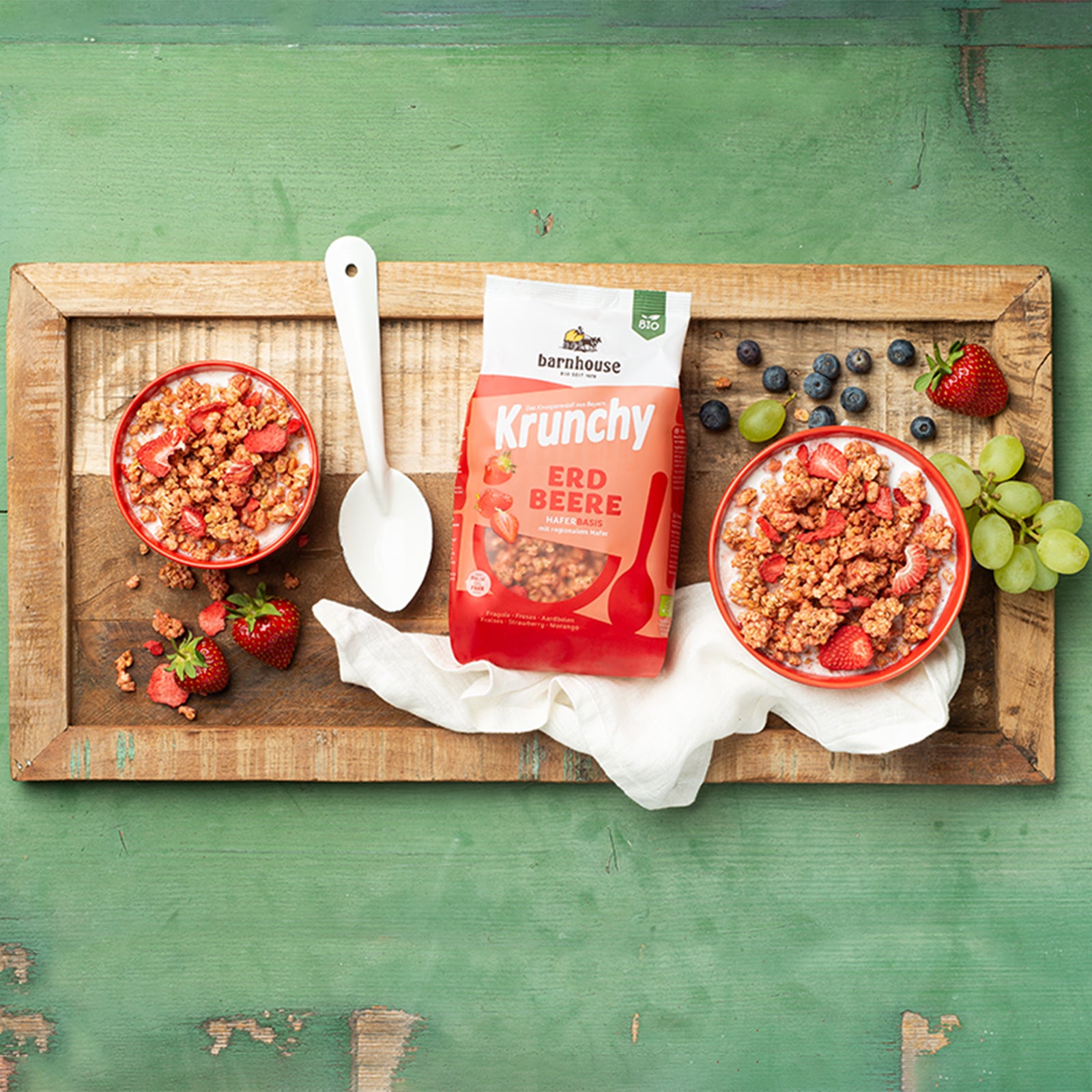 Handgemachtes Holzbrett, das auf einem grün angestrichenen Holztisch liegt, der schon etwa abblättert. In der Mitte liegt ein Beutel Krunchy Erdbeere von Bio-Knuspermüsli-Hersteller Barnhouse, darunter ein weißes Tuch und daneben ein großer, weißer Löffel. Außerdem stehen zwei Müslischüsseln auf dem Brett, die mit Milch und Krunchy Erdbeere befüllt sind. Für einen noch schöneren Anblick sorgen Müslicluster, Erdbeerscheiben, frische Erdbeeren, Heidelbeeren und Weintrauben, die auf dem Holzbrett verteilt sind