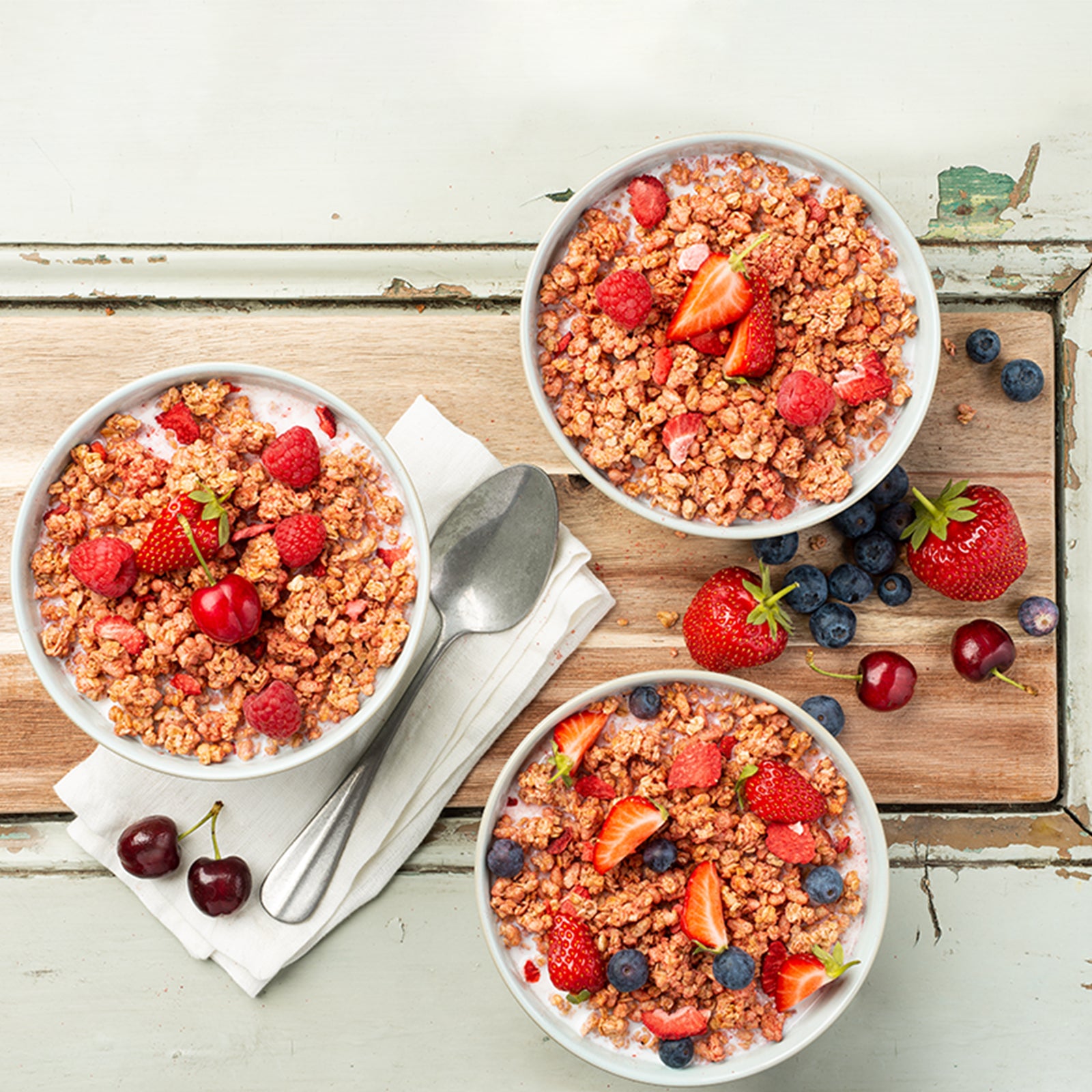 Blick von oben auf ein etwas abgenutztes Holzbrett, auf dem drei Müslischalen mit Krunchy Erdbeere von Bio-Knuspermüsli-Hersteller Barnhouse, Milch und frischen Erdbeeren, Himbeeren, Heidelbeeren und einer Kirsche stehen. Außerdem liegen noch eine weiße Serviette und ein silberner Löffel daneben. Für einen noch schöneren Anblick sorgen frische Erdbeeren, Heidelbeeren, Kirschen und ein paar Müsli-Cluster.