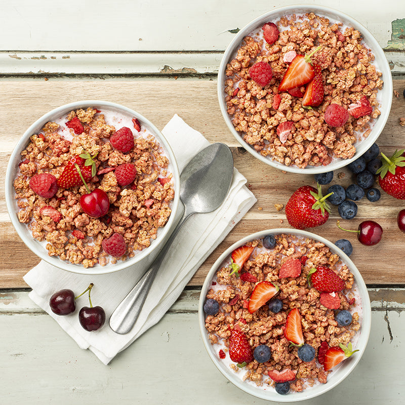 Blick von oben auf ein etwas abgenutztes Holzbrett, auf dem drei Müslischalen mit Krunchy Erdbeere von Bio-Knuspermüsli-Hersteller Barnhouse, Milch und frischen Erdbeeren, Himbeeren, Heidelbeeren und einer Kirsche stehen. Außerdem liegen noch eine weiße Serviette und ein silberner Löffel daneben. Für einen noch schöneren Anblick sorgen frische Erdbeeren, Heidelbeeren, Kirschen und ein paar Müsli-Cluster.