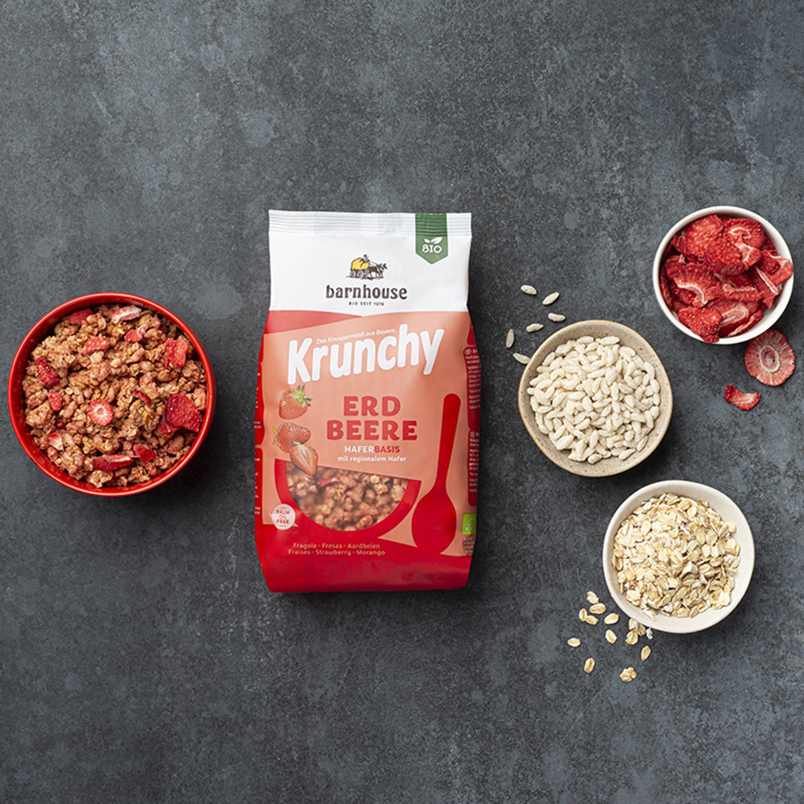 Beutel Krunchy Erdbeere von Bio-Knuspermüsli-Hersteller Barnhouse auf schwarzer Marmorplatte. Außenrum verschiedenfarbige Schüsseln mit dem besagten Müsli sowie dessen Zutaten Haferflocken, Reispops, und Erdbeerscheiben. Die Zutaten liegen auch noch teilweise neben den Schüsseln verteilt auf dem Tisch.