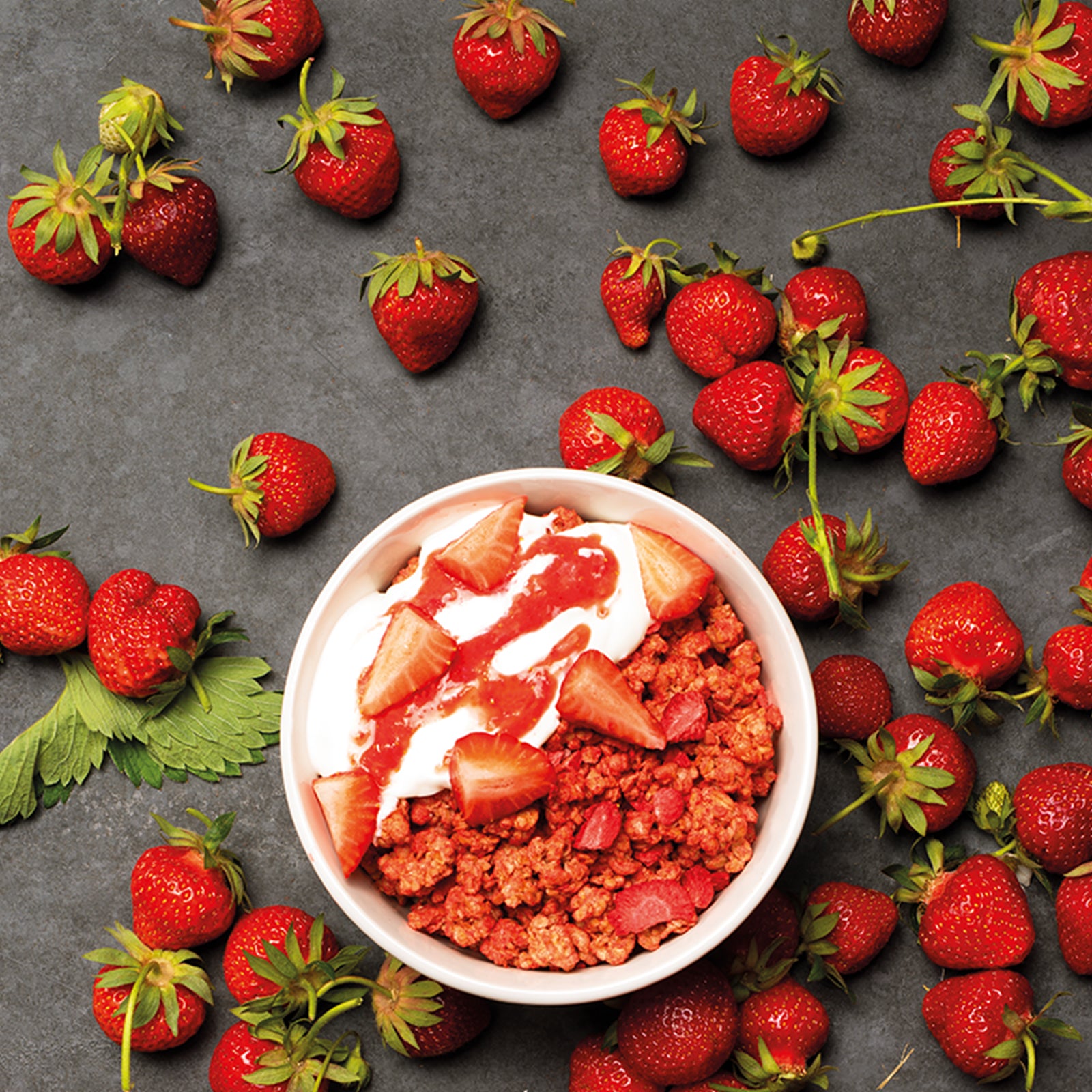 Jede Menge frische Erdbeeren, die auf einem schwarzen Marmor-Untergrund liegen. In der Mitte steht eine weiße Schale mit Krunchy Erdbeere von Bio-Knuspermüsli-Hersteller Barnhouse, Joghurt, Erdbeercreme und frischen, geschnitten Erdbeerstücken.
