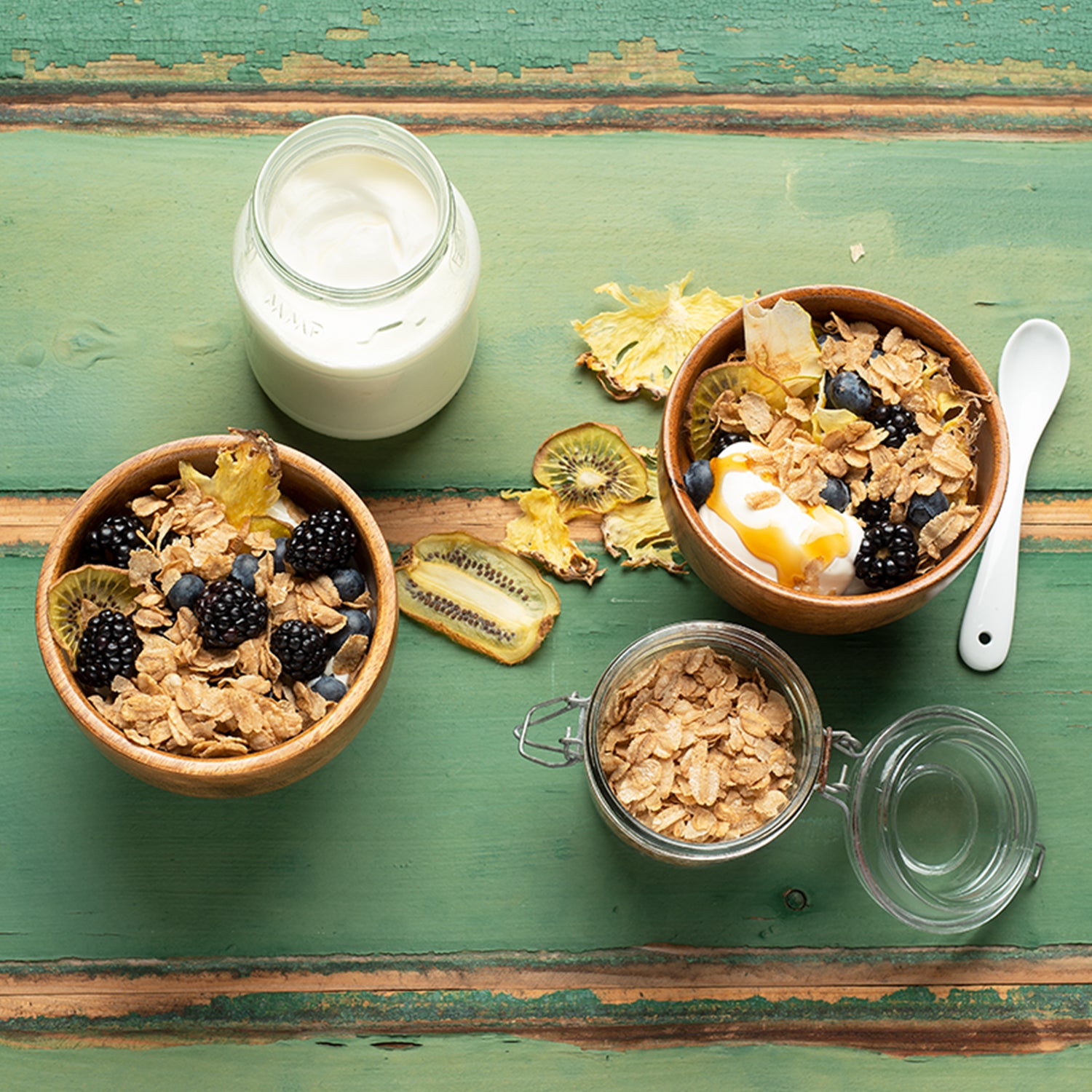 Holztisch, der grün gestrichen ist und schon an einigen Stellen abblättert. Darauf befinden sich 2 hohe Schalen gefüllt mit Dinkelflakes, Kiwi und Brombeeren. In der einen Schale sind noch Joghurt und Honig mit drin. Außerdem befinden sich noch ein Gefäß rein mit Flakes Dinkel von Bio-Knuspermüsli-Hersteller Barnhouse, eine Kanne mit Joghurt, ein weißer Löffel sowie Kiwi- und weitere Obstscheiben auf dem Tisch.
