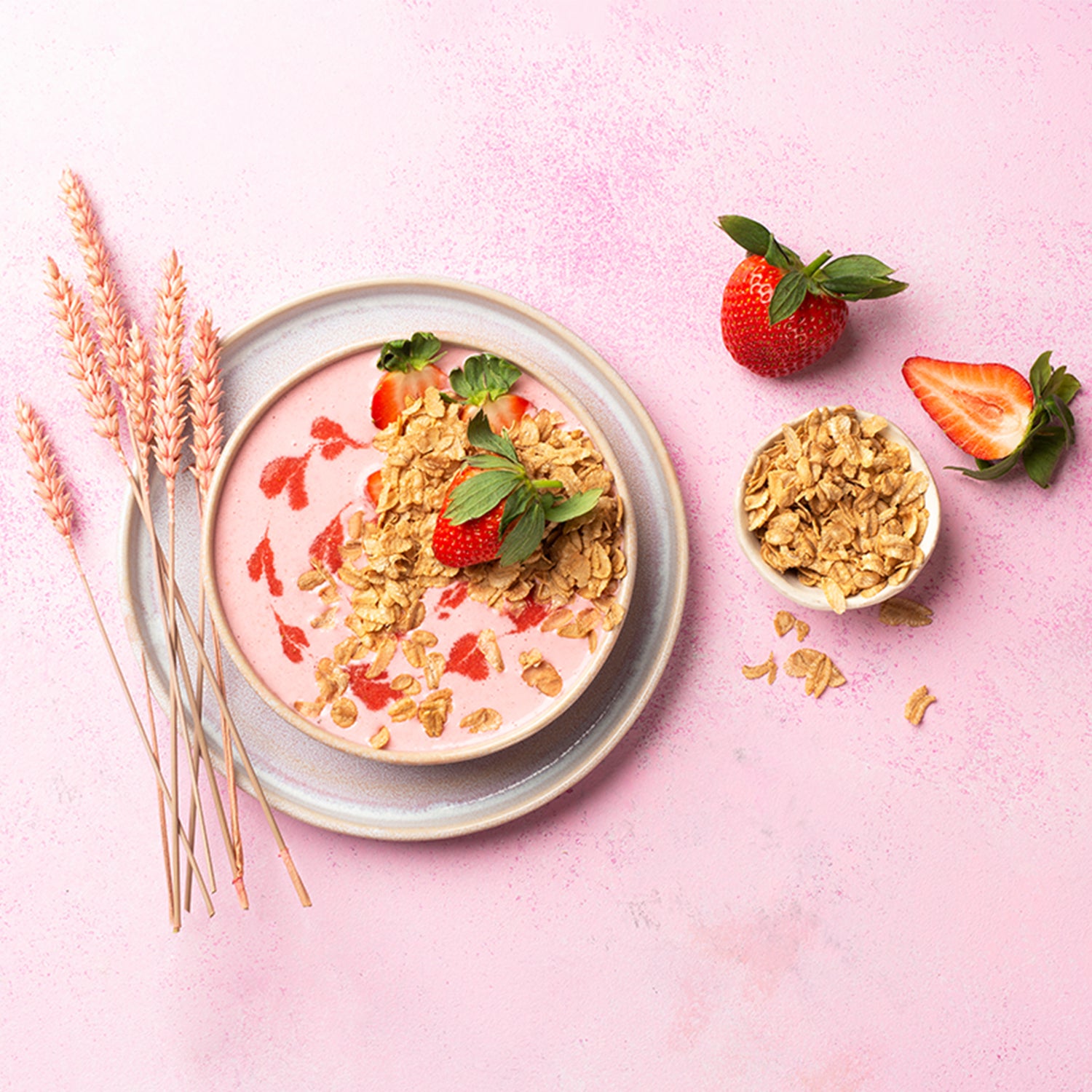 Rosa Untergrund, auf dem ein grauer Teller steht. Auf diesem Teller befindet sich eine Schüssel, die mit rosa Joghurt, Erdbeeren und Flakes Dinkel vom Bio-Knuspermüsli-Hersteller Barnhouse gefüllt ist. Neben der Schüssel befinden sich Dinkel-Halme sowie eine kleine Schüssel mit Dinkelflakes. Außerdem liegen noch 2 Erdbeeren sowie weitere Dinkelflakes auf dem Tisch.