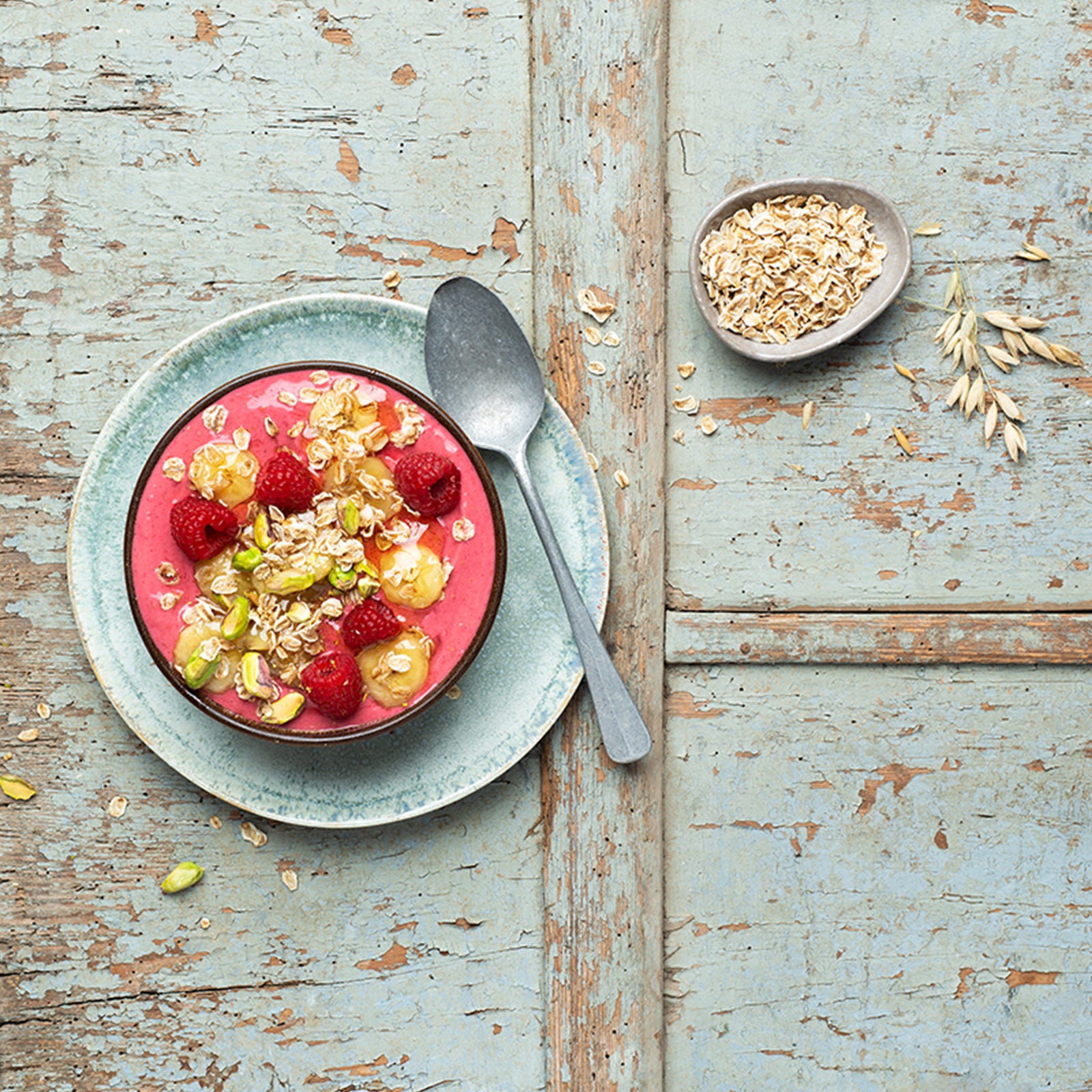 Holztisch, der blau-gräuilch gestrichen ist und schon an einigen Stellen abblättert. Darauf ein blauer Teller mit Schüssel mit rötlichem Joghurt, Himbeeren, Bananenscheiben und Pistazien. Als Topping sind dort noch Flakes Hafer von Bio-Knuspermüsli-Hersteller Barnhouse mit drauf. Auf dem Teller liegt noch ein großer, grauer Löffel. Außerdem liegen noch zwei Haferhalme sowie Pistazien und vereinzelte Haferflocken auf dem Tisch. Rechts oben steht noch eine kleine, graue Schüssel rein mit Haferflakes.