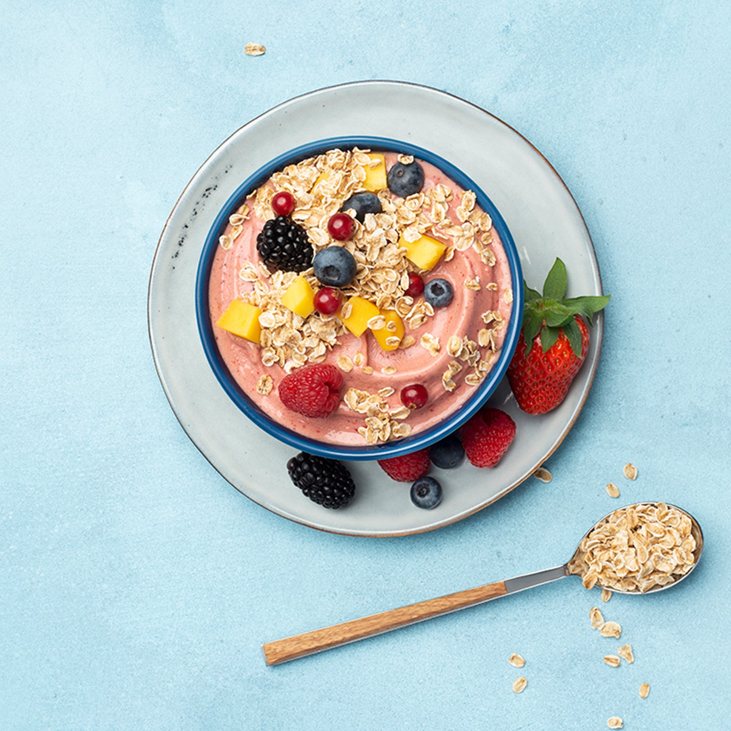 Hellblauer Untergrund, auf dem ein weiß-grauer Teller steht. Auf diesem Teller befindet sich eine dunkelblaue Schüssel, die mit rötlichem Joghurt, jeder Menge Beeren und Mango befüllt ist. Auch auf dem Unterteller befinden sich noch verschiedene Beeren. Als Topping sind noch Flakes Hafer von Bio-Knuspermüsli-Hersteller Barnhouse mit drauf.. Neben der Schüssel befindet sich ein grau-brauner Löffel, der mit Haferflakes gefüllt ist. Daneben liegen noch weitere Haferflakes.