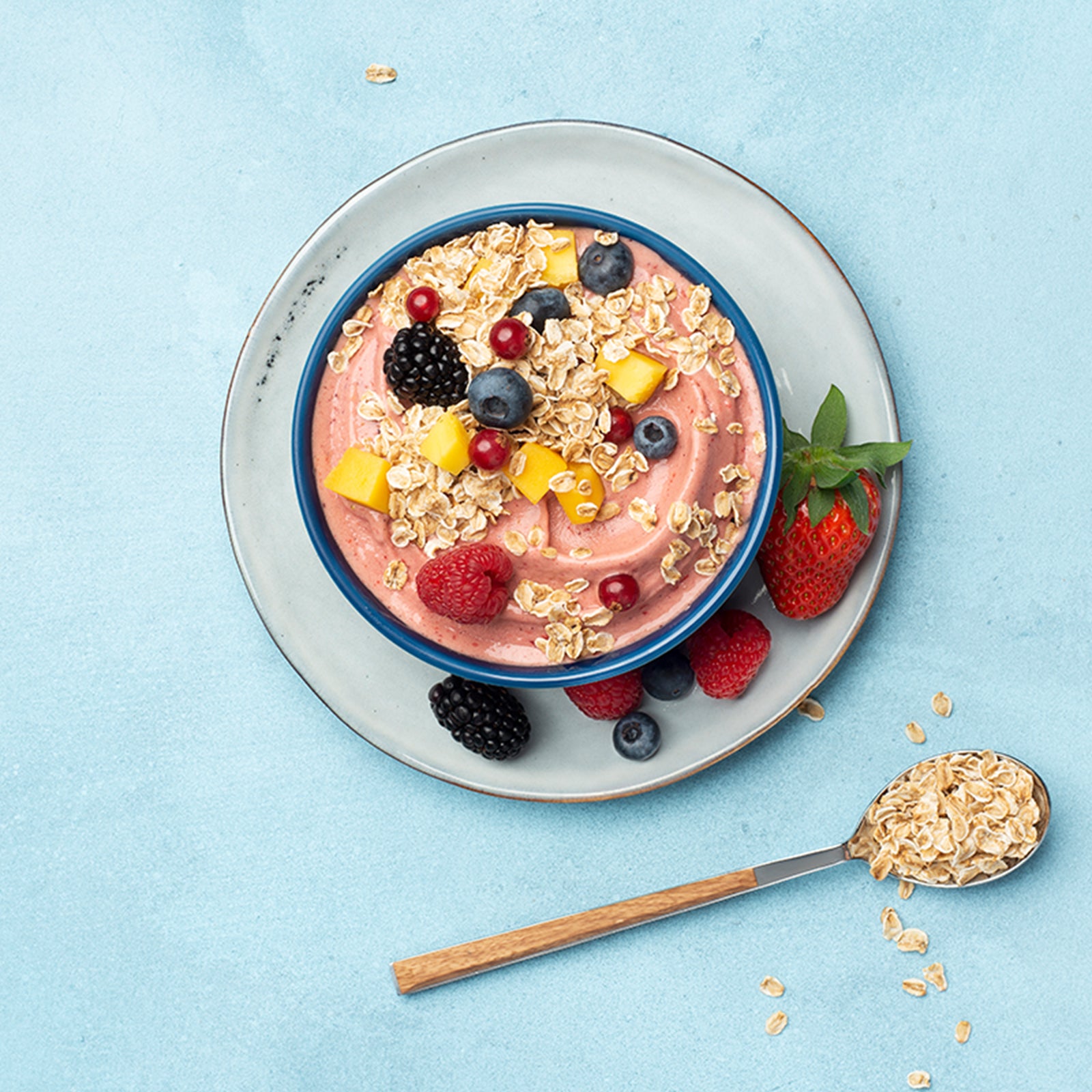 Hellblauer Untergrund, auf dem ein weiß-grauer Teller steht. Auf diesem Teller befindet sich eine dunkelblaue Schüssel, die mit rötlichem Joghurt, jeder Menge Beeren und Mango befüllt ist. Auch auf dem Unterteller befinden sich noch verschiedene Beeren. Als Topping sind noch Flakes Hafer von Bio-Knuspermüsli-Hersteller Barnhouse mit drauf.. Neben der Schüssel befindet sich ein grau-brauner Löffel, der mit Haferflakes gefüllt ist. Daneben liegen noch weitere Haferflakes.