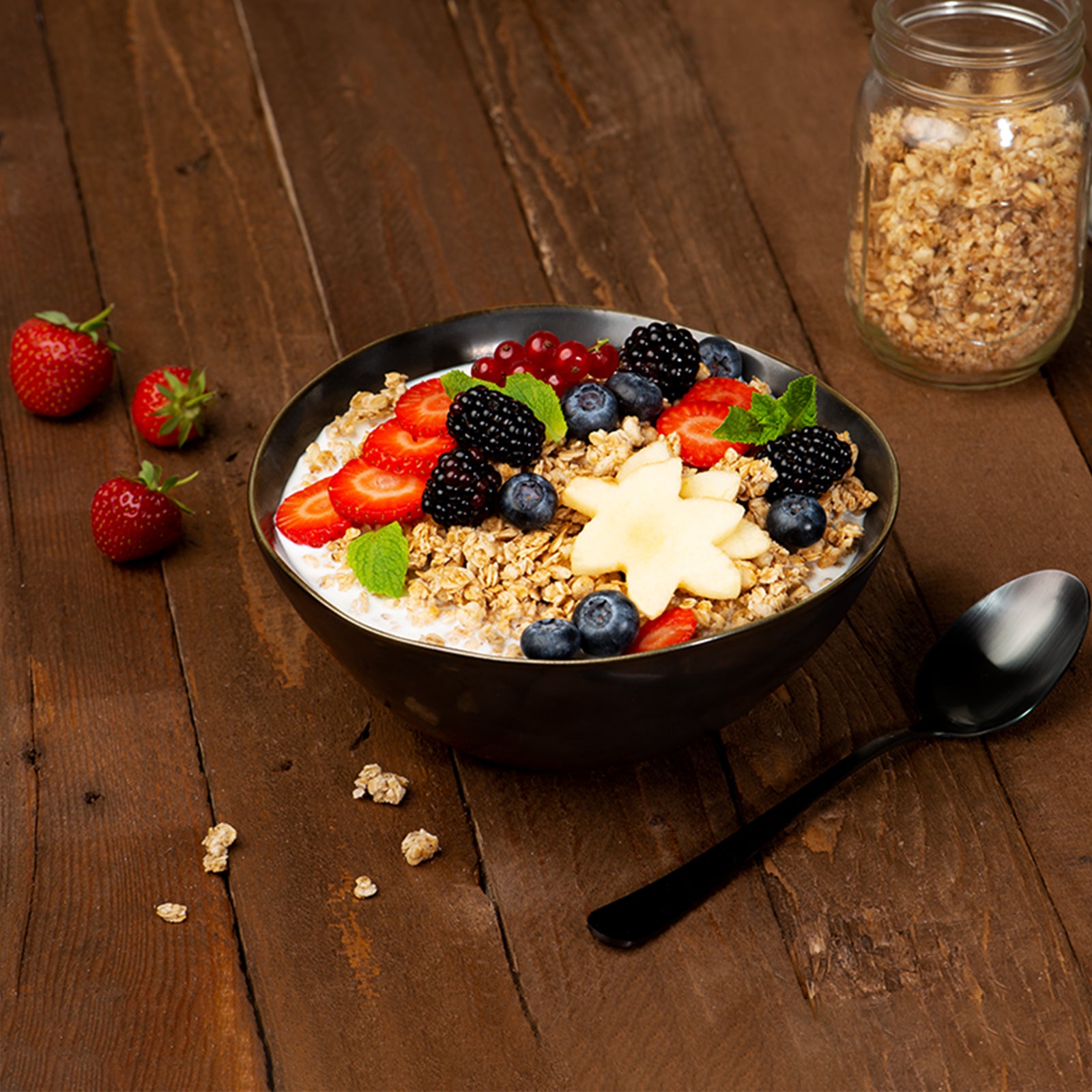 Eine sehr schön angerichtete Bowl mit Krunchy Hafer alternativ gesüßt, frischen Brombeeren, Erdbeerscheiben, Johannisbeeren, Heidelbeeren, Sternenfrucht und etwas Minze. Die schwarze Müslischüssel steht auf einem Holztisch, auf dem noch frische Erdbeeren, ein paar Müslicluster und ein Löffel liegen. Im Hintergrund ist noch ein durchsichtiges Glas mit dem Hafer-Müsli zu sehen.