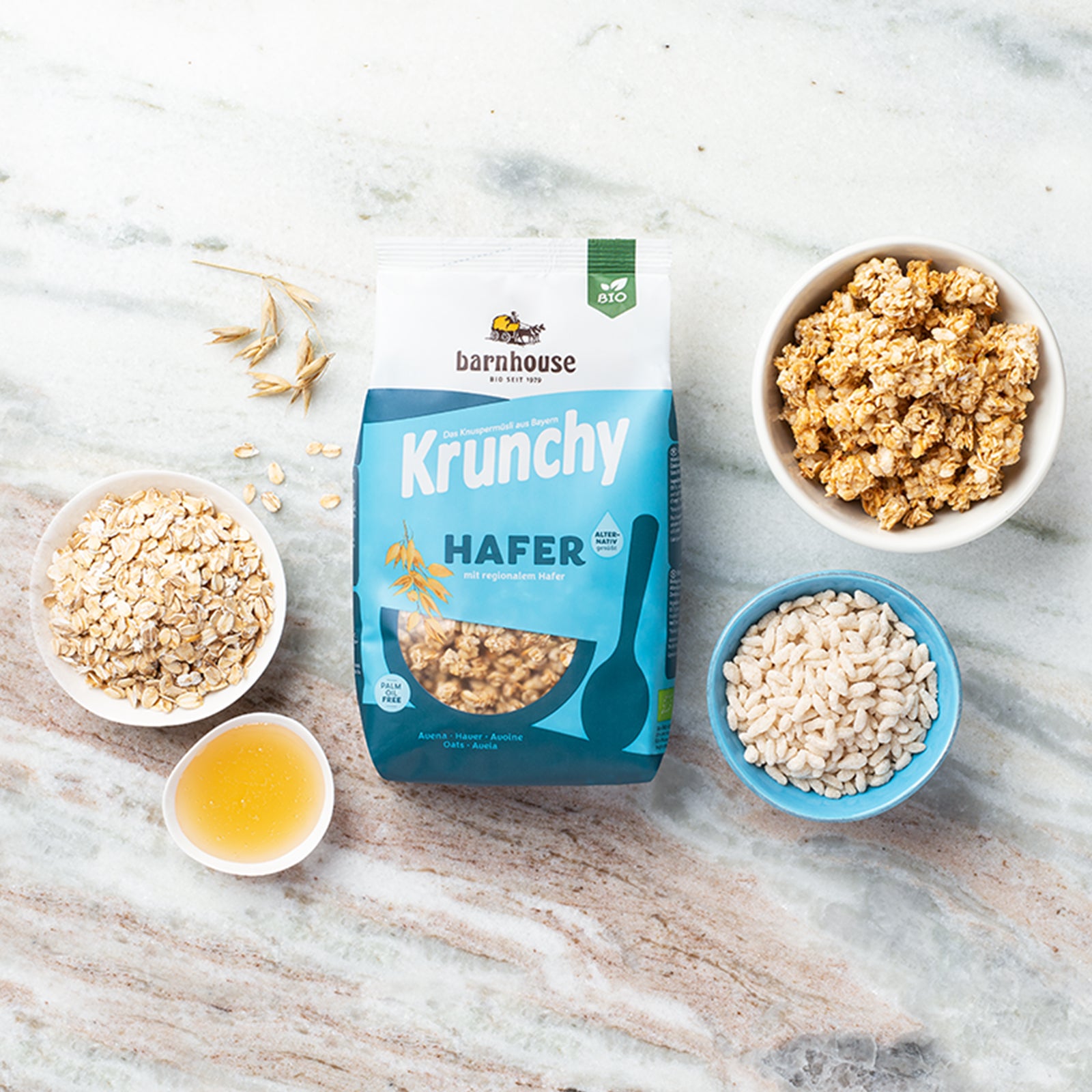 Beutel Krunchy Hafer alternativ gesüßt von Bio-Knuspermüsli-Hersteller Barnhouse auf heller Marmorplatte. Daneben steht eine weiße Schale mit diesem Müsli. Zudem stehen noch zwei weiße Schalen mit den Zutaten Haferflocken sowie Sonnenblumenöl sowie eine hellblaue Schale mit gepufftem Hafer daneben. Auf dem Tisch sind noch ein paar Haferflocken und -knospen verteilt.