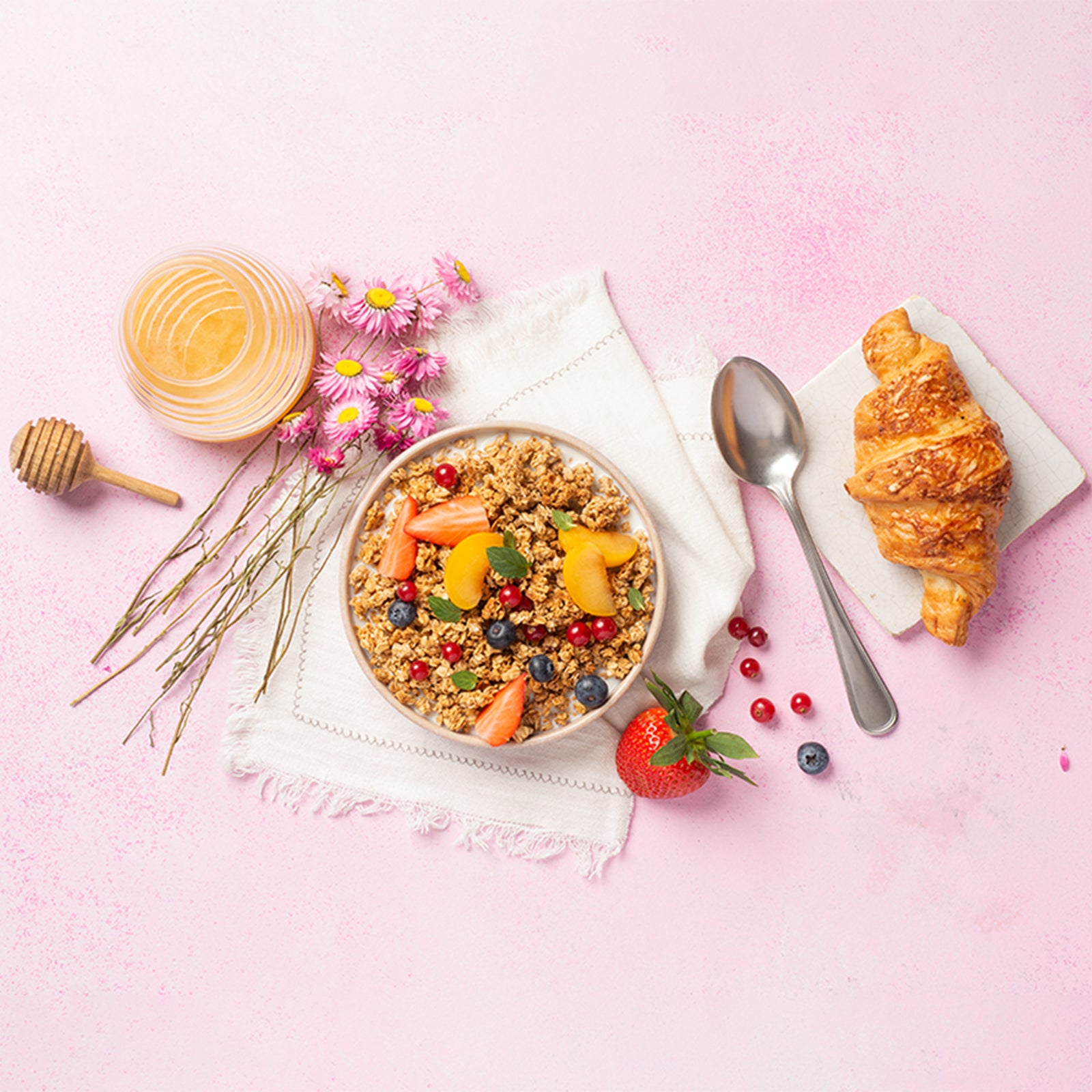 Wunderbar angerichtete Frühstücksbowl mit Krunchy Honig und einem Topping aus verschiedenen, frischen Früchten und Obst. Die weiße Schale steht auf einer weißen Decke und ist umgeben von einem Glas Honig, einem Honiglöffel, rosa Blumen, einer frischen Erdbeere, Heidelbeere und Johannisbeeren. Am rechten Bildrand liegt noch ein Löffel sowie ein frisches Schinken-Käse-Croissant auf einer Serviette. Der Untergrund ist eine Mischung aus Weiß und Rosa.