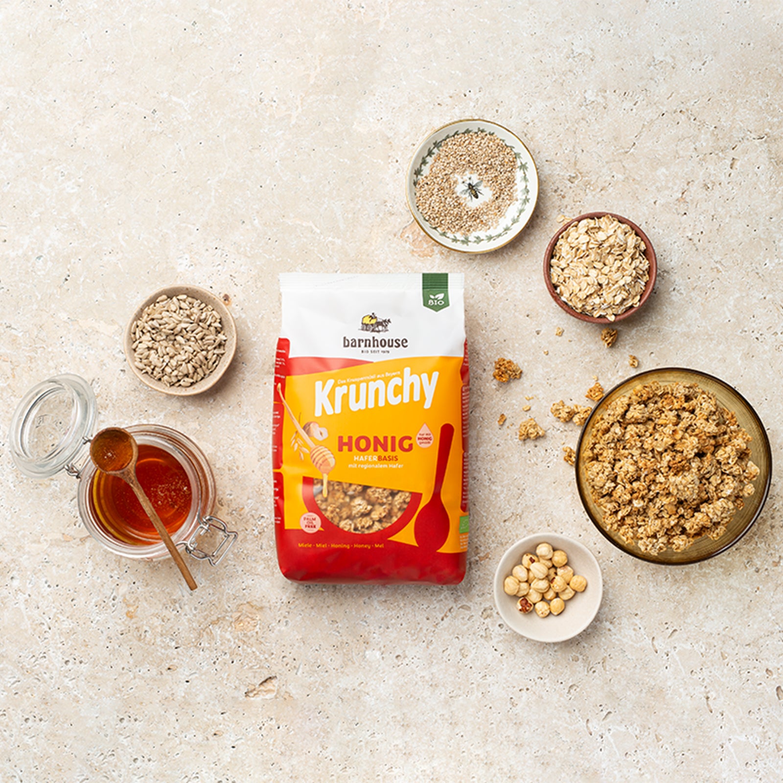 Beutel Krunchy Honig von Bio-Knuspermüsli-Hersteller Barnhouse auf heller Marmorplatte. Außenrum steht eine bräunliche Schale mit dem Krunchy-Honig sowie verschiedenfarbige Schüsseln mit den Zutaten Honig, Sesam, Sonnenblumenkerne, Haselnüsse und Haferflocken. Auf dem Tisch sind noch ein paar Cluster des Müslis verteilt.