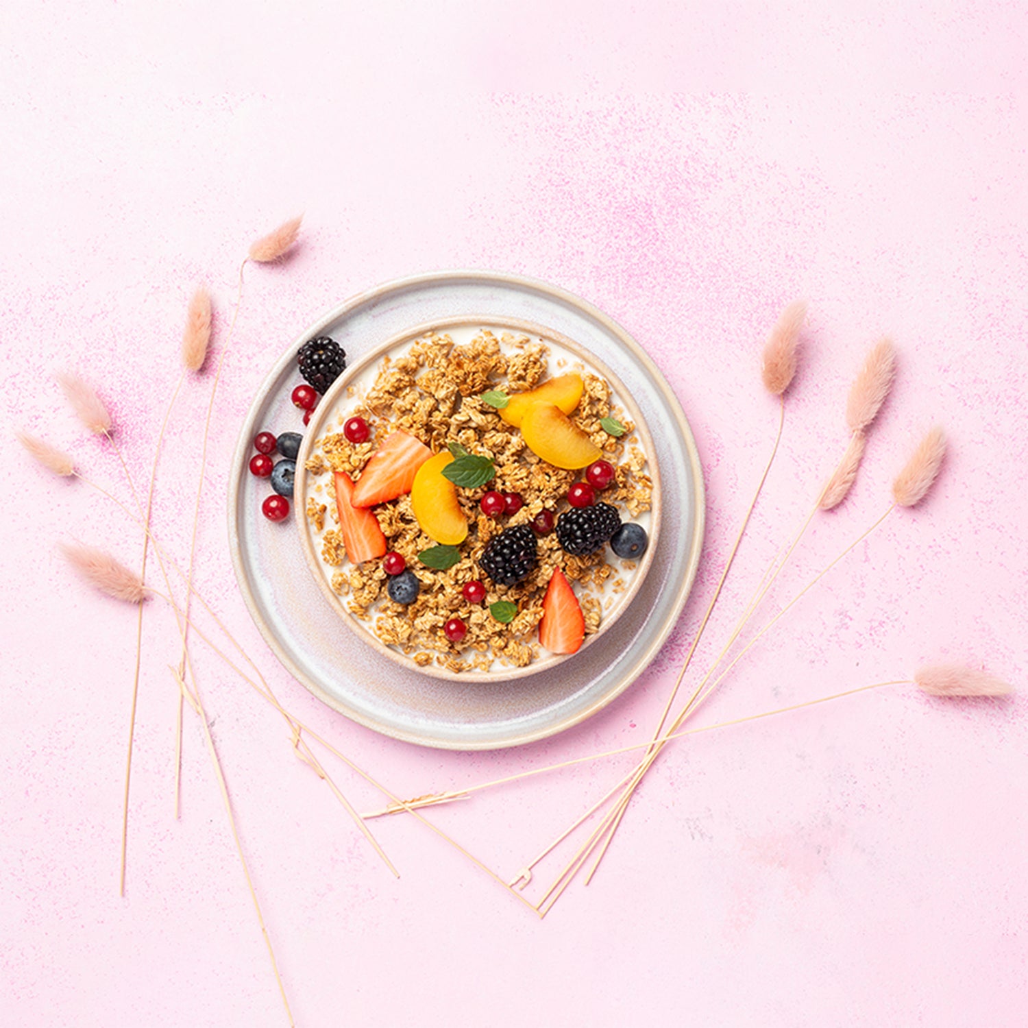 Rosa Untergrund mit flauschigen Blumen, auf dem ein weißer Unterteller mit Johannisbeeren, Heidelbeeren und einer Brombeere steht. Darauf befindet sich eine Schüssel mit Milch, Krunchy Honig, frischen Erdbeerstücken, Aprikosen, Johannisbeeren, Heidelbeeren, Brombeeren und etwas Minze.
