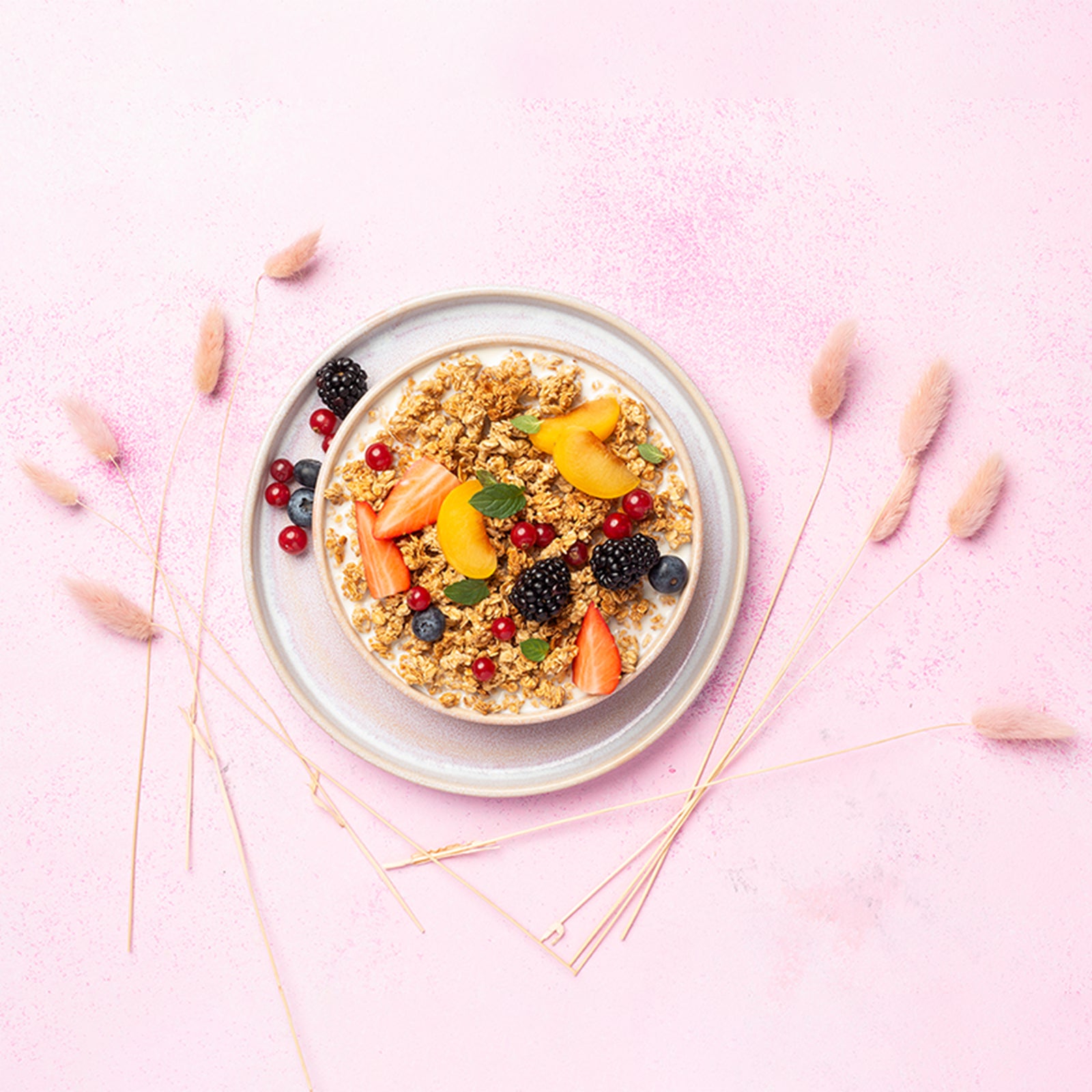 Rosa Untergrund mit flauschigen Blumen, auf dem ein weißer Unterteller mit Johannisbeeren, Heidelbeeren und einer Brombeere steht. Darauf befindet sich eine Schüssel mit Milch, Krunchy Honig, frischen Erdbeerstücken, Aprikosen, Johannisbeeren, Heidelbeeren, Brombeeren und etwas Minze.