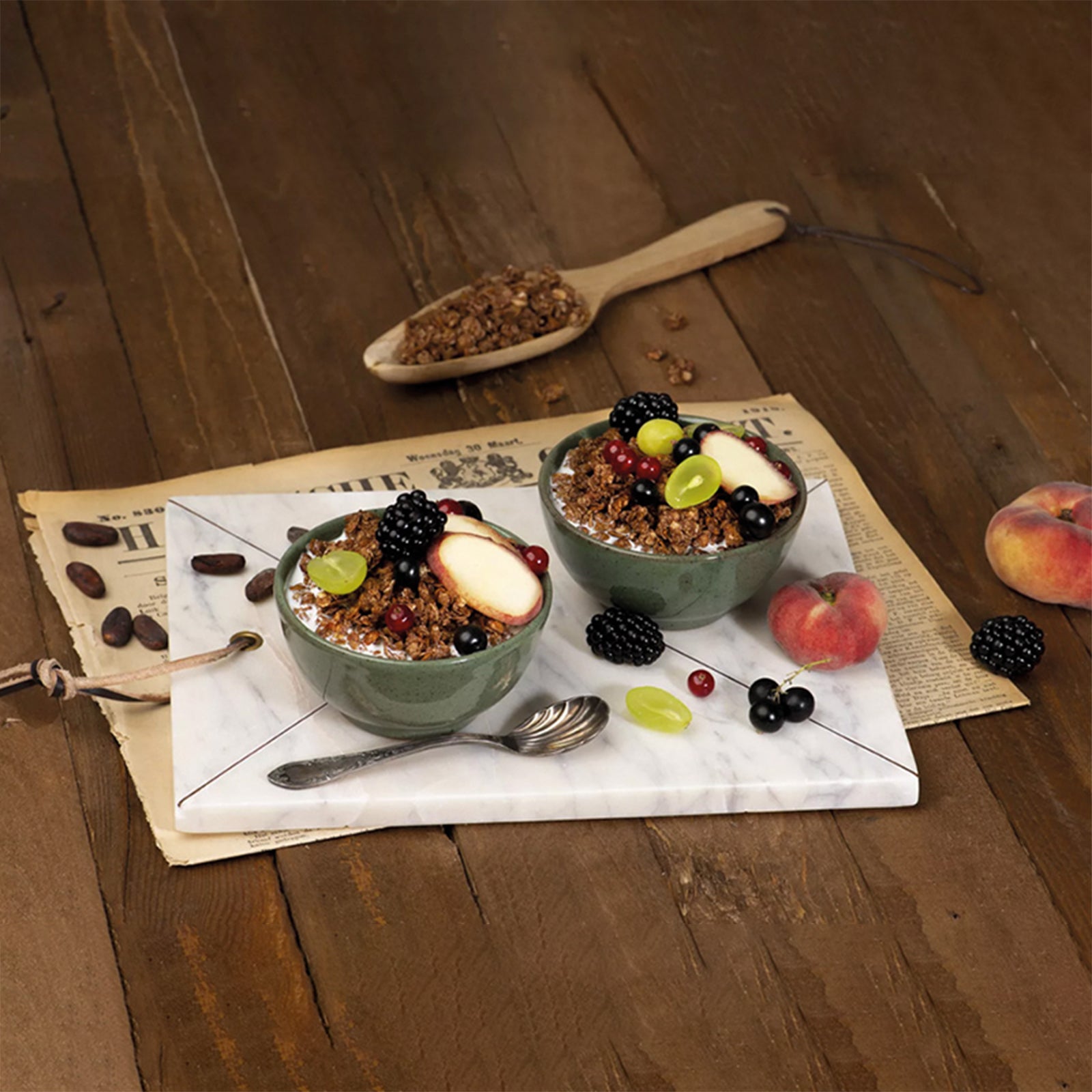 2 grüne Müslischalen, die mit Milch und Krunchy Joy Cocoa von Bio-Knuspermüsli-Hersteller Barnhouse befüllt und mit Weintrauben, Apfelscheiben, Johannisbeeren und Brombeeren schön dekoriert sind. Darunter eine weiße Steinplatte mit prunkvollem Löffel sowie den besagten Beeren, einer Aprikose und ein paar Kaffeebohnen. Darunter liegt eine sehr alte Zeitung, auf der wiederum ein paar Kaffeebohnen liegen. Der Untergrund ist ein brauner Holztisch, auf dem ein alter Holzlöffel mit dem besagten Müsli liegt.