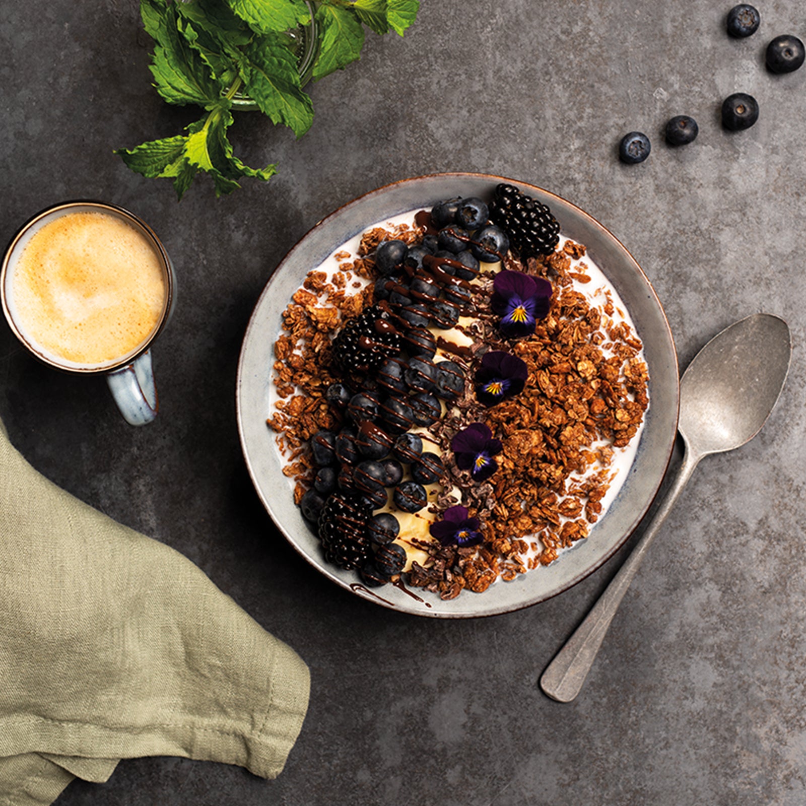Graue Marmorplatte, auf der Minzblätter, eine Tasse Cappuccino, Heidelbeeren, ein grün-graues Geschirrtuch und ein Löffel zu sehen sind. Den Mittelpunkt stellt eine graue Müslischüssel da, die mit etwas Milch und Krunchy Joy Cocoa von Bio-Knuspermüsli-Hersteller Barnhouse befüllt ist. Als Topping liegen Heidelbeeren und Brombeeren obendrauf, die mit einer Schokosoße betröpfelt sind. Als besonderes Highlight wurden noch vier essbare, violette Blumen auf dem Müsli platziert.
