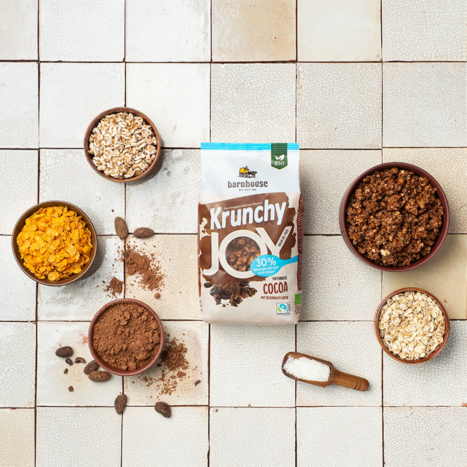 Beutel Krunchy Joy Cocoa von Bio-Knuspermüsli-Hersteller Barnhouse auf gemusterten Fließen. Darunter liegt noch eine braune Holzschaufel mit Meersalz. Außerdem stehen außenrum noch vier braune Schalen mit den Zutaten Haferflocken, Cornflakes, gepuffter Dinkel und Kakaopulver sowie eine braune Schale mit dem zuckerreduzierten Müsli. Kakaopulver und -bohnen sind auf dem Tisch verteilt.
