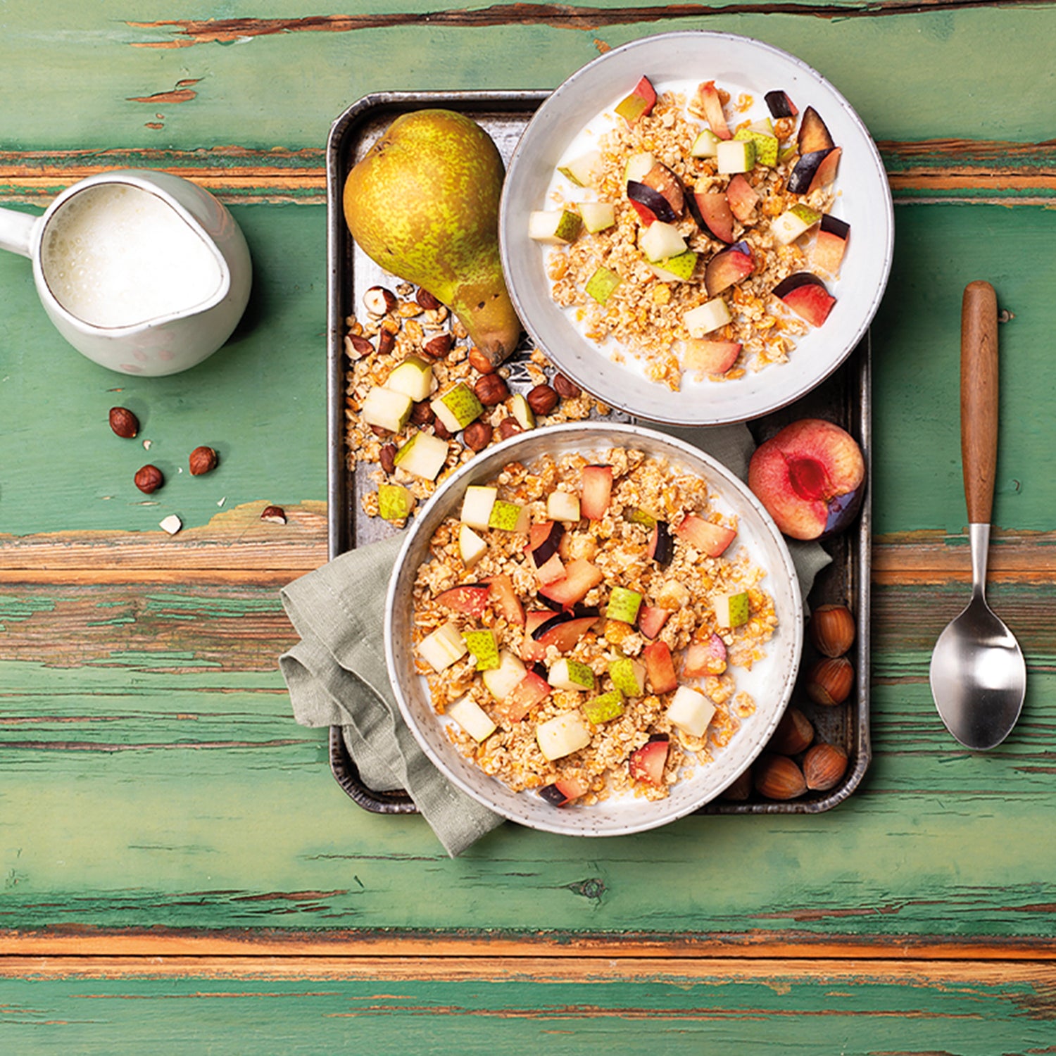 Blick von oben auf 2 weiße Müslischalen, die mit Milch und Krunchy Joy Nuss von Knuspermüsli-Hersteller Barnhouse befüllt und mit bunter Obst- und Fruchtauswahl schön dekoriert sind. Die beiden Schalen stehen auf einem silbernen Tablett, auf dem eine halbe Pflaume, eine ganze Birne, weitere Birnenstücke sowie Haselnüsse und Knuspermüsli-Cluster liegen. Auf dem grün gestrichenen Holztisch liegt noch ein silbernen Löffel mit Holzgriff und neben einer kleine Karaffe mit Milch liegen paar Haselnüsse.