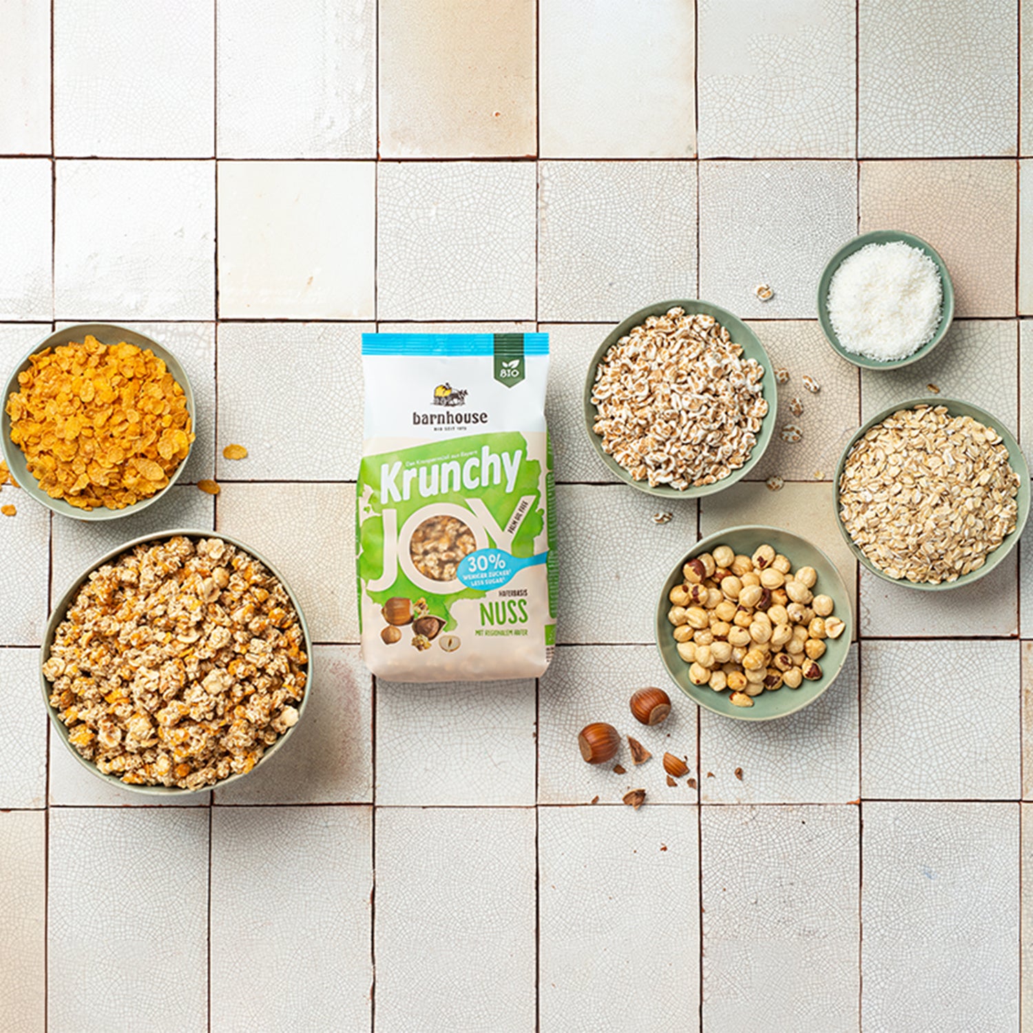 Grün-weißer Beutel Krunchy Joy Nuss von Bio-Knuspermüsli-Hersteller Barnhouse auf gemusterten Fließen. Außenrum stehen fünf grüne Schalen mit den Zutaten Cornflakes, gepuffter Dinkel, Kokosraspeln, Haselnüsse und Haferflocken.  Die Zutaten liegen auch um die Schalen herum verteilt. Eine weitere grüne Schale ist mit dem nussigen, zuckerreduzierten Knuspermüsli befüllt.