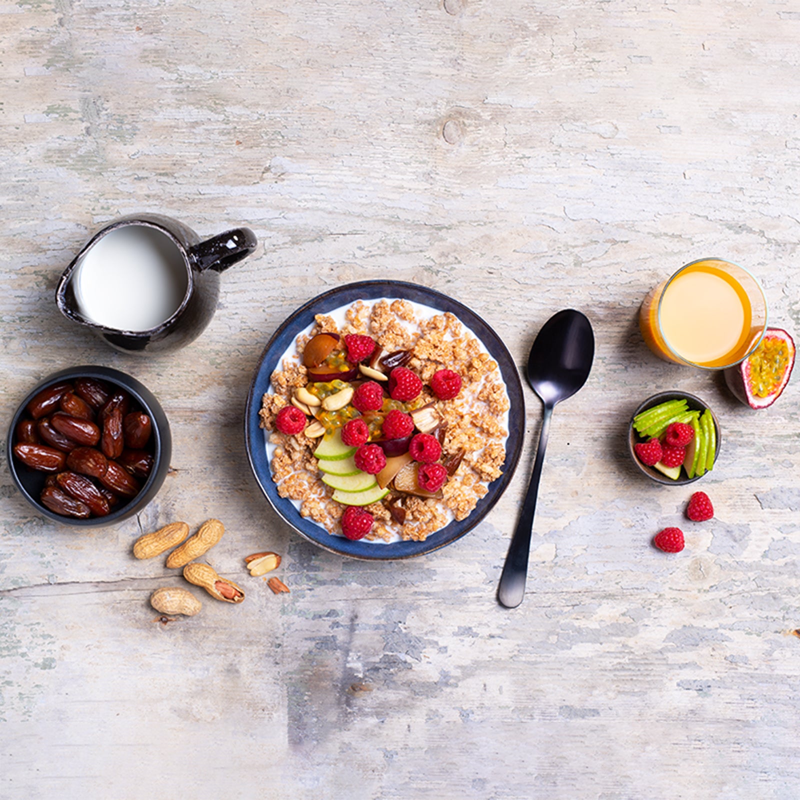 Ein heller Holztisch, in dessen Mitte eine blaue Müslischale steht, die mit Milch & Krunchy Klassik Hafer von Bio-Knuspermüsli-Hersteller Barnhouse befüllt ist. Als Topping liegen Himbeeren, Apfelscheiben, Nüsse, Pflaumen, Datteln und eine Passionsfrucht schön angerichtet obendrauf. Rechts daneben sind ein Löffel, ein Glas Orangensaft, eine halbe Passionsfrucht, eine Schüssel mit Apfelscheiben und Himbeeren. Links steht eine schwarze Kanne mit Milch, eine schwarze Schale mit Datteln und Erdnüsse liegen da.