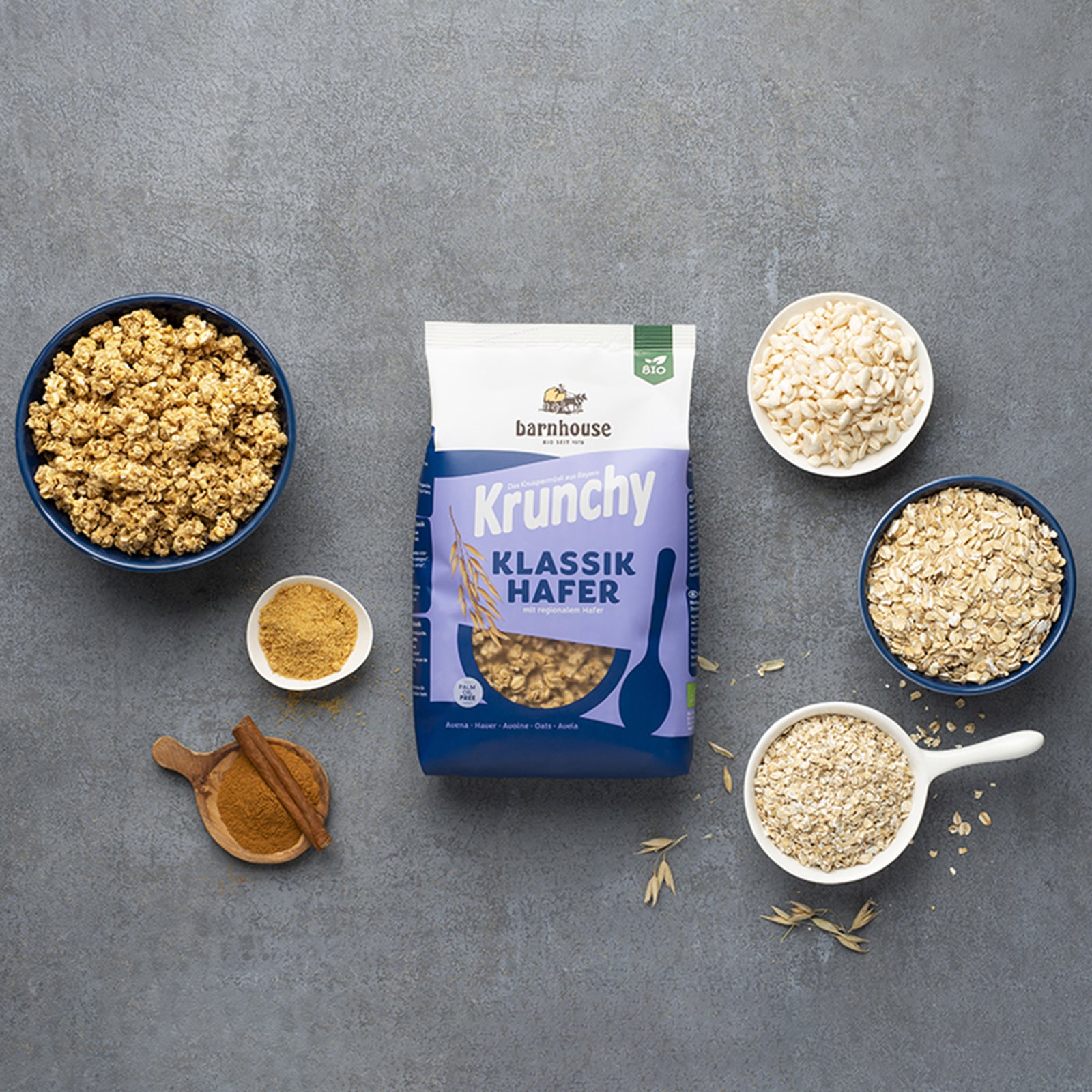 Blau-weißer Beutel Krunchy Klassiker Hafer von Bio-Knuspermüsli-Hersteller Barnhouse auf dunkelgrauem Marmortisch. Außenrum stehen verschiedenfarbige Schalen mit den Zutaten Haferflocken, gepuffter Reis, Vollruhrzucker und gemahlenem Zimt inkl. Zimtstange. Die Zutaten liegen auch um die Schalen herum verteilt. Eine weitere Schale ist mit dem Knuspermüsli befüllt.