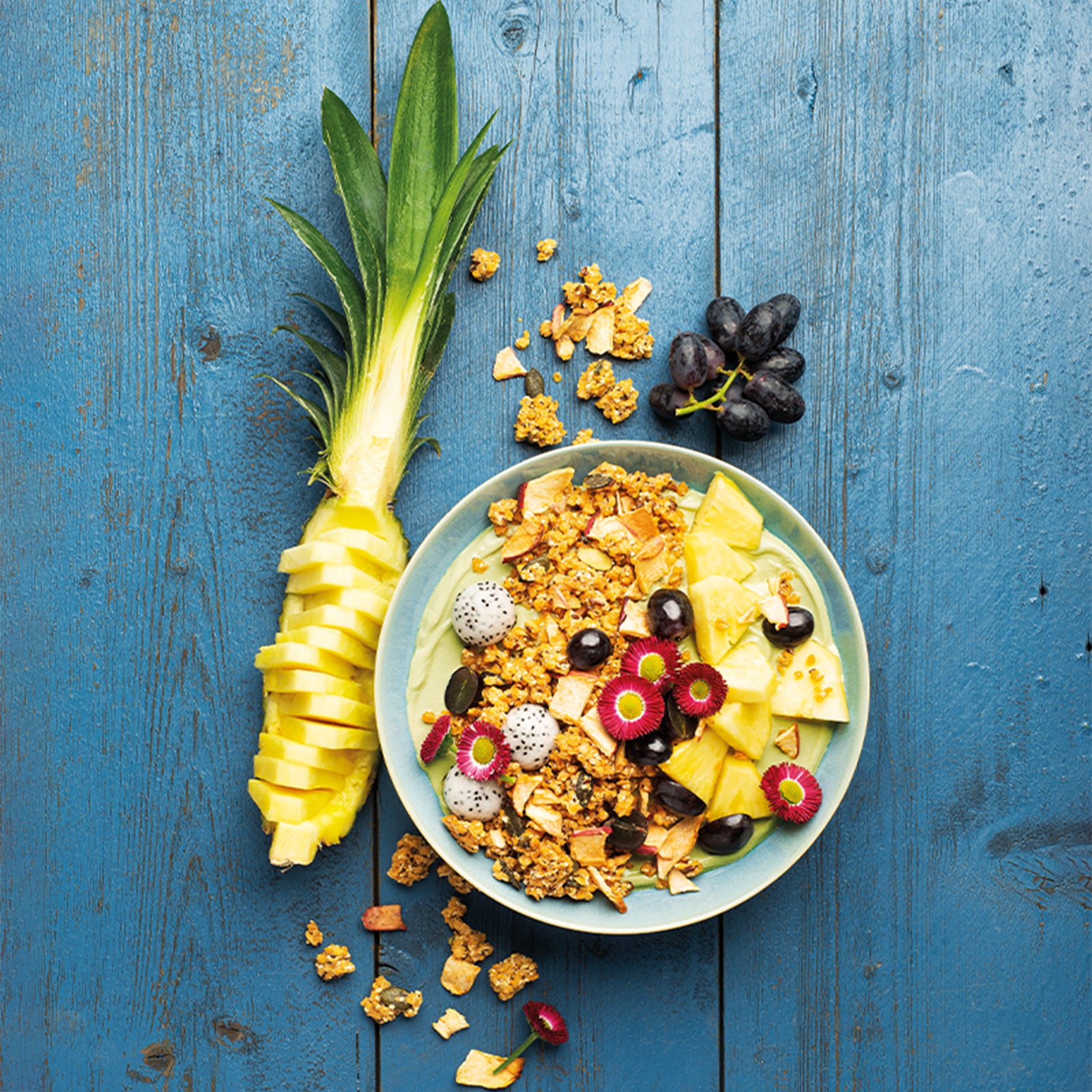 Blauer Holztisch, in dessen Mitte eine hellblaue Müslischüssel mit fruchtigem Joghurt und Krunchy Linse mit Apfel von Bio-Knuspermüsli-Hersteller Barnhouse steht. Für einen besonderen Anblick des Müslis sorgt das Topping aus Ananas, blauen Weintrauben, Sternenfrucht-Kugeln und essbaren, roten Blüten. Neben der Schale liegt eine aufgeschnittene Ananas, ein paar blaue Weintrauben sowie etwas Linsen-Müsli und eine rote, essbare Blüte.