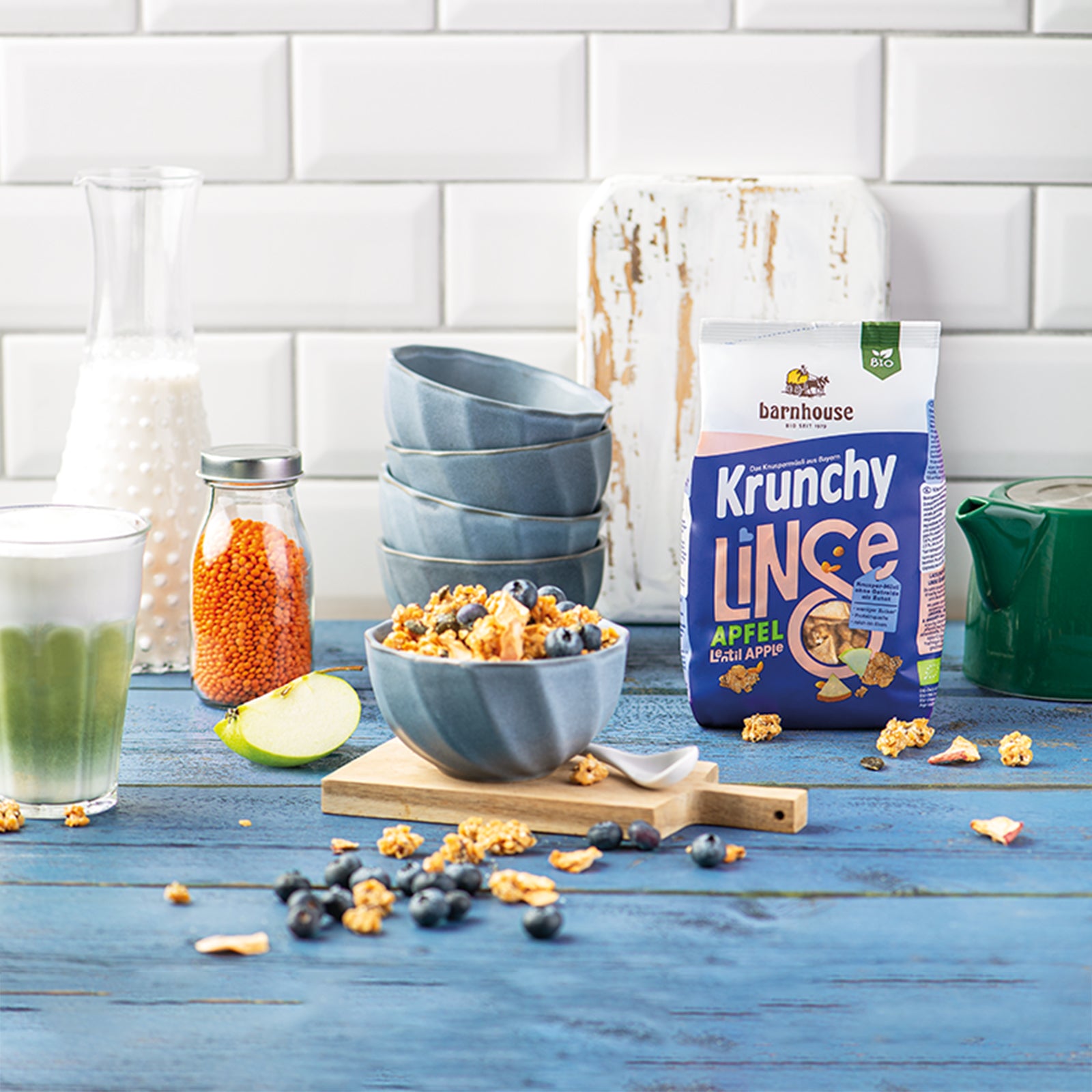 Küchenzeile mit blauer Holzablage & weißer Ziegelwand. In der Mitte eine blaue Müslischüssel mit Krunchy Linse Apfel von Bio-Knuspermüsli-Hersteller Barnhouse auf einem Holzbrett mit weißem Löffel. Auf der Holzablage sind noch einige Müsli-Cluster, Heidelbeeren, Apfelstücke und Sonnenblumenkerne verteilt. Im Hintergrund steht ein Glas mit Apfel-Smoothie sowie eine Karaffe mit Milch und ein Gefäß mit roten Linsen. Im Hintergrund noch 4 weitere, gestapelte Müslischalen, 1 Schneidebrett & 1 Kaffeekanne.