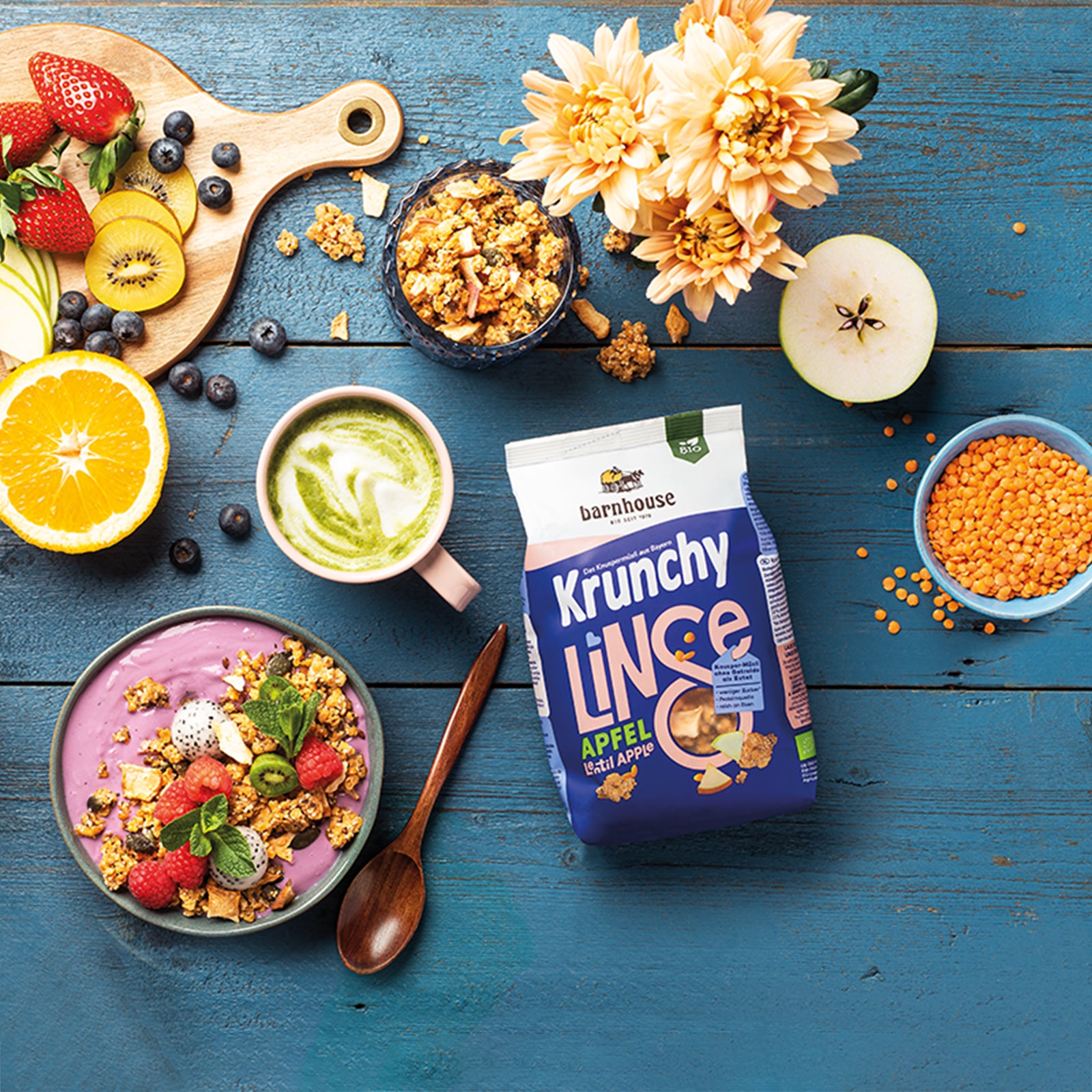 Sehr schön dekorierter, blauer Holztisch mit einem weiß-blauen Beutel Krunchy Linse mit Apfel von Bio-Knuspermüsli-Hersteller Barnhouse. Das Müsli findet sich auch in einem Glas sowie einer blauen Schale mit Beeren-Joghurt, Himbeeren, Kiwi, Sternenfrucht und Minze wieder. Ein Matcha Latte, eine blaue Schale mit roten Linsen, eine cremefarbene Blume, ein Holzlöffel,eine Holzbrett mit frischen Erdbeeren, Apfelscheiben, Kiwis & Heidelbeeren sowie eine Orange & ein Apfel sorgen für einen noch schöneren Anblick.