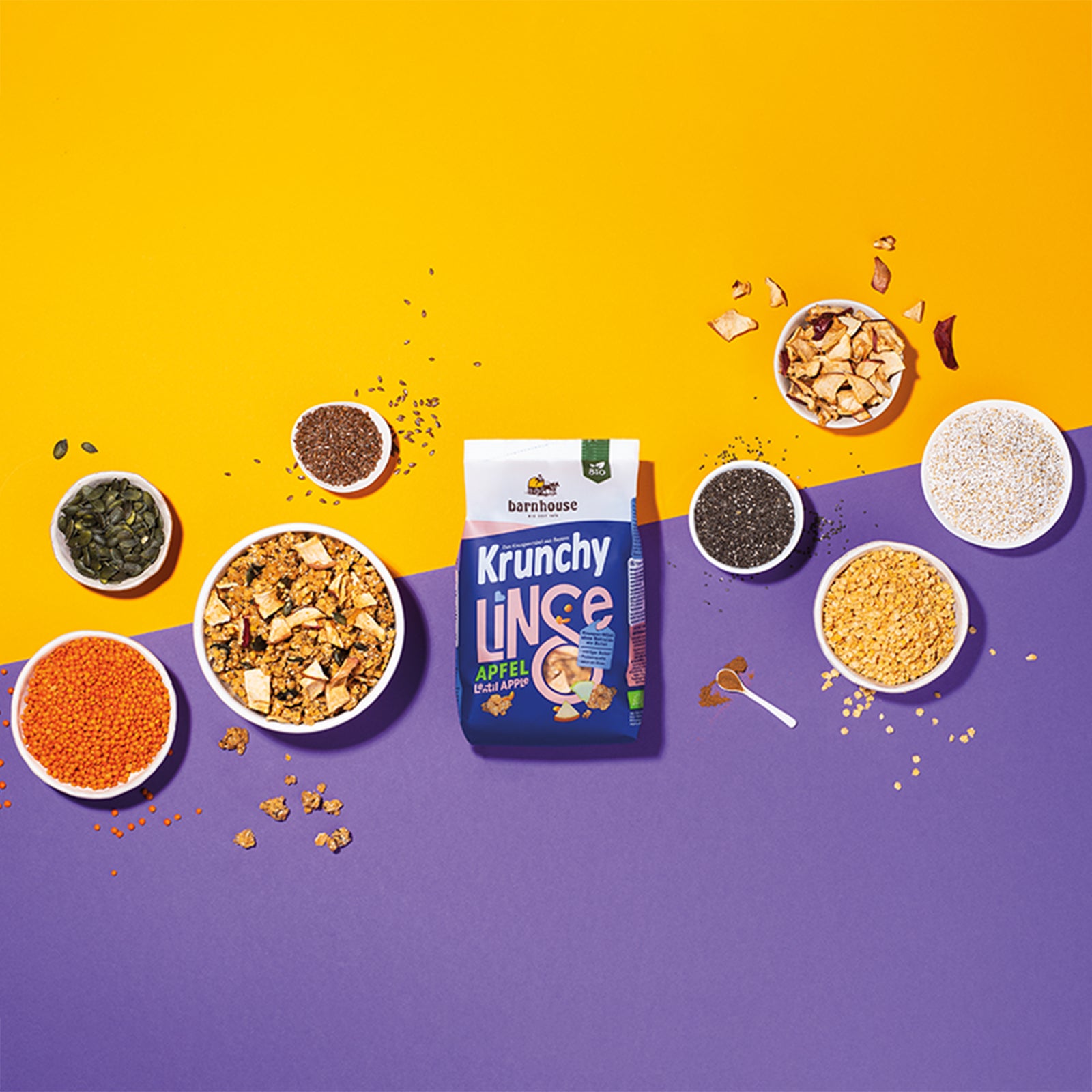 Zweifarbiger Hintergrund mit oranger Farbe oben und violetter Farbe unten. In der Mitte liegt ein blau-weißer Beutel Krunchy Linse mit Apfel von Bio-Knuspermüsli-Hersteller Barnhouse. Außenrum stehen sieben weiße Schalen mit den Zutaten rote Linsen, Kichererbsenmehl, gepuffter Amaranth , Leinsamen, Chiasamen, Kürbiskerne und getrocknete Apfelstücke sowie ein weißer Löffel mit Zimt. Die Zutaten liegen auch um die Schalen herum verteilt. Eine weitere weiße Schale ist mit dem Linsen-Müsli befüllt.