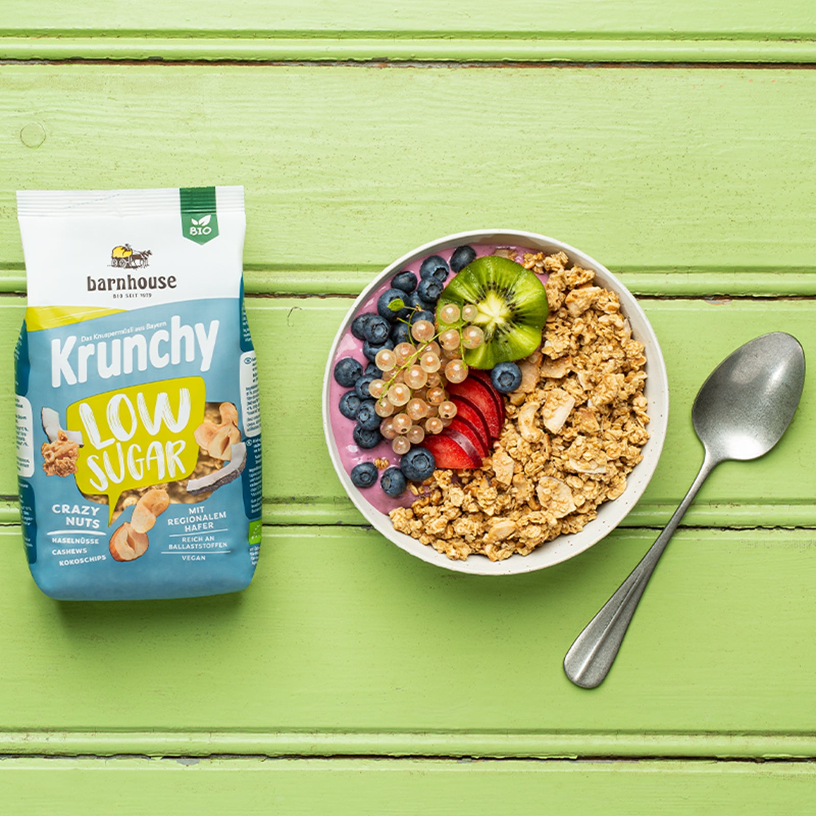 Ein wundervoll angerichtete Müsli-Bowl mit beerigem Joghurt und Krunchy Low Sugar Crazy Nuts von Bio-Knuspermüsli-Hersteller Barnhouse. Für ein optisches Highlight sorgt das Topping aus weißen Johannisbeeren, Heidelbeeren, einer Kiwi in Blütenform und ein paar Scheiben Pflaumen. Auf dem grün angestrichenen Holztisch liegt noch ein silberner Löffel und ein geschlossener, grün-weiß-blauer Beutel des Müslis.