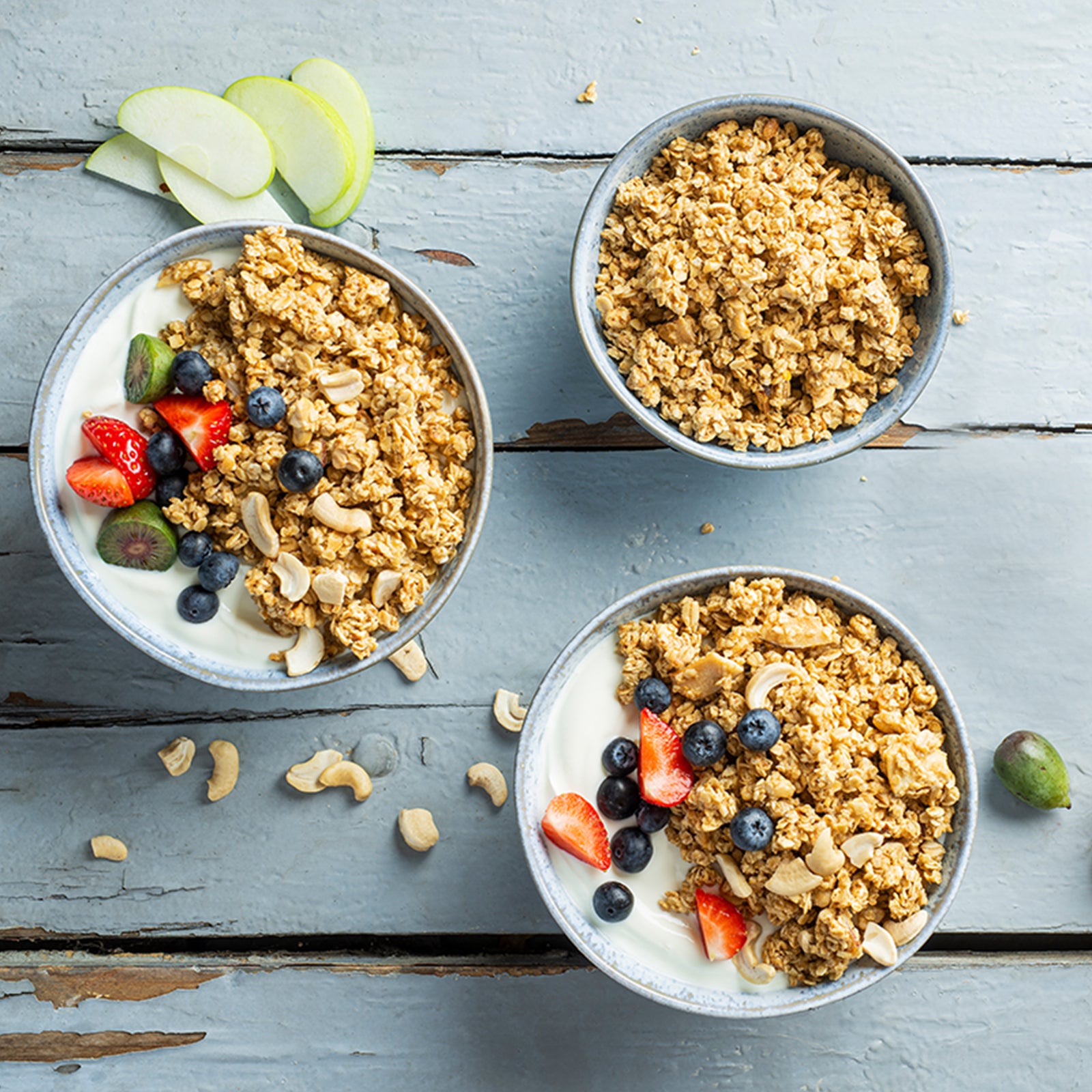 Blick von oben auf einen Holztisch, auf dem ein paar grüne Apfelspalten, eine Erdbeere, eine Minikiwi und Cashewnüsse liegen. Oben rechts steht eine blaue Müslischüssel mit Krunchy Low Sugar Crazy Nuts von Bio-Knuspermüsli-Hersteller Barnhouse. Im unteren Bereich steht eine weitere blaue Schale mit dem zuckerarmen Nuss-Müsli, allerdings mit Joghurt und Erdbeeren sowie Heidelbeeren als Topping. Links steht noch eine blaue Müslischale, die zudem noch mit zwei halben Minikiwis dekoriert ist.
