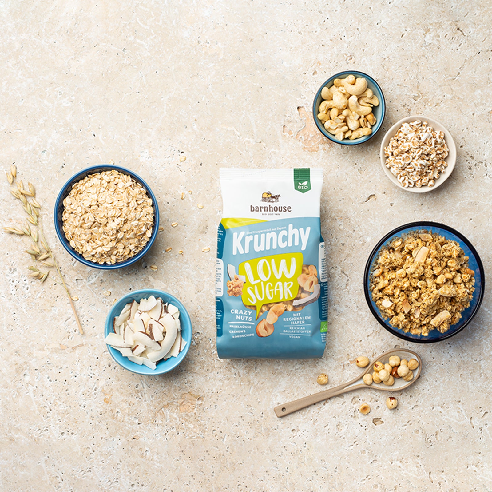 Blick von oben auf einen grün-weiß-blauen Beutel Krunchy Low Sugar Crazy Nuts von Bio-Knuspermüsli-Hersteller Barnhouse auf einer beigen Marmorplatte. Außenrum stehen vier verschiedenfarbige Schalen mit den Zutaten Kokoschips, Cashewnüssen, gepuffter Dinkel & Haferflocken sowie ein Löffel mit Haselnüssen. Eine weitere Schale ist mit dem nussigen, zuckerarmen Knuspermüsli befüllt. Ein Haferhalm liegt auch dabei.