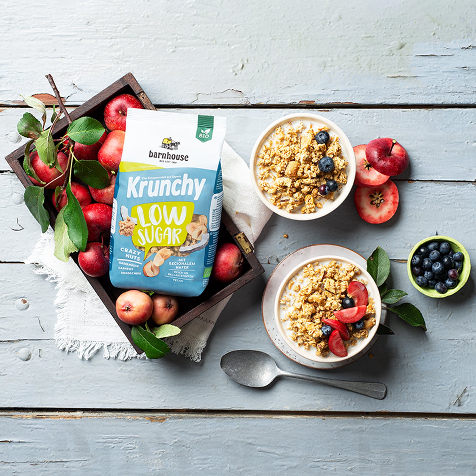 Blick von oben auf einen grün-weiß-blauen Beutel Krunchy Low Sugar Crazy Nuts von Bio-Knuspermüsli-Hersteller Barnhouse, der in einer Kiste mit roten Äpfeln auf einer Decke steht. Daneben stehen 2 weiße Schüsseln mit Milch und diesem zuckerarmen Müsli, jeweils mit Heidelbeeren dekoriert. Eine Schale steht auf einem Unterteller und enthält zusätzlich Erdbeerscheiben. Auf dem gräulich gestrichenen Holztisch liegen ein silberner Löffel, eine grüne Schale mit Heidelbeeren, ein aufgeschnittener Apfel & Blätter.