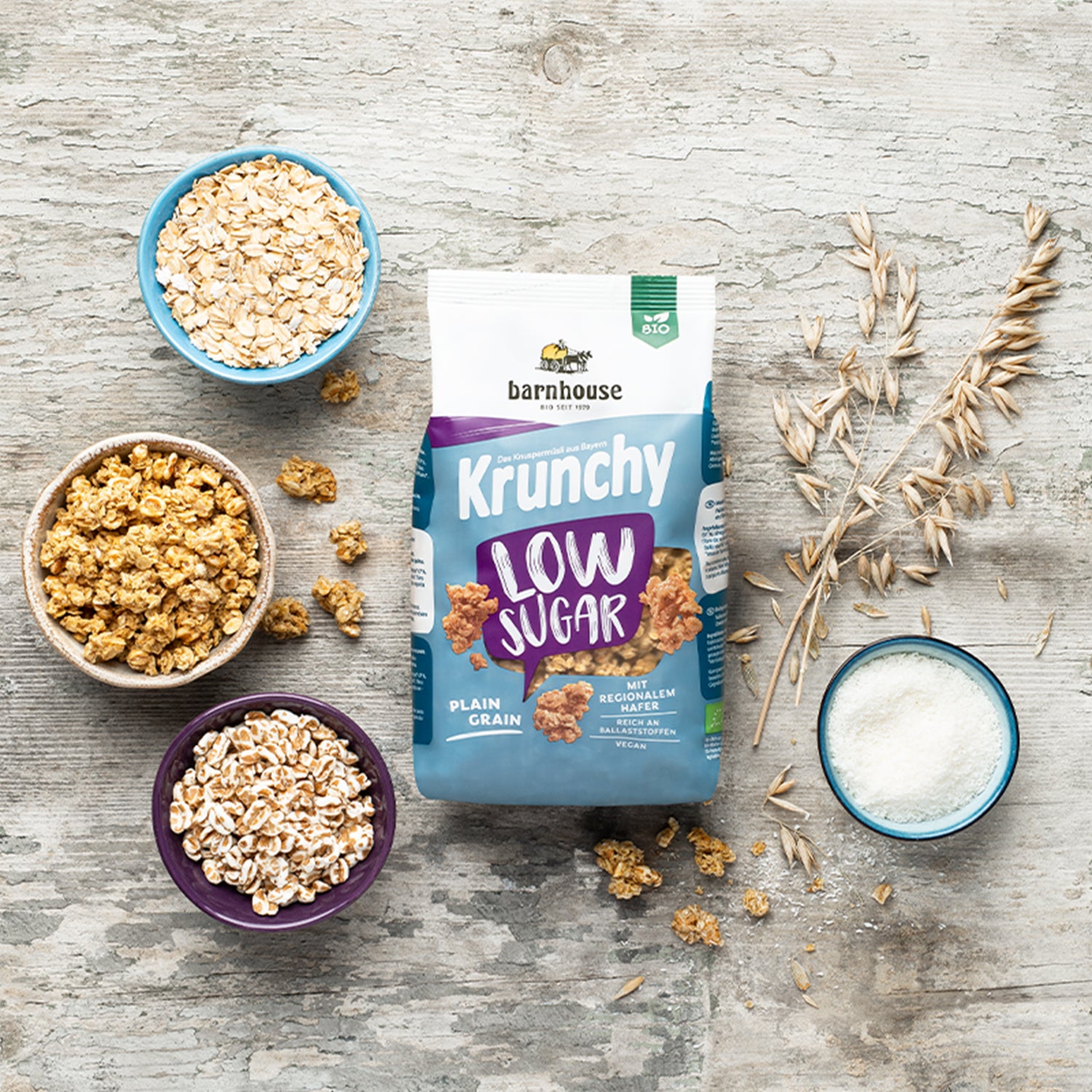 Blick von oben auf einen lila-weiß-blauen Beutel Krunchy Low Sugar Plain Grain von Bio-Knuspermüsli-Hersteller Barnhouse. Außenrum stehen drei verschiedenfarbige Schalen mit den Zutaten gepuffter Dinkel, Kokosraspeln und Haferflocken. Eine weitere Schale ist mit dem zuckerarmen Knuspermüsli befüllt. Auf dem Holztisch liegen außerdem noch ein Haferhalm sowie Müsli-Cluster verteilt.