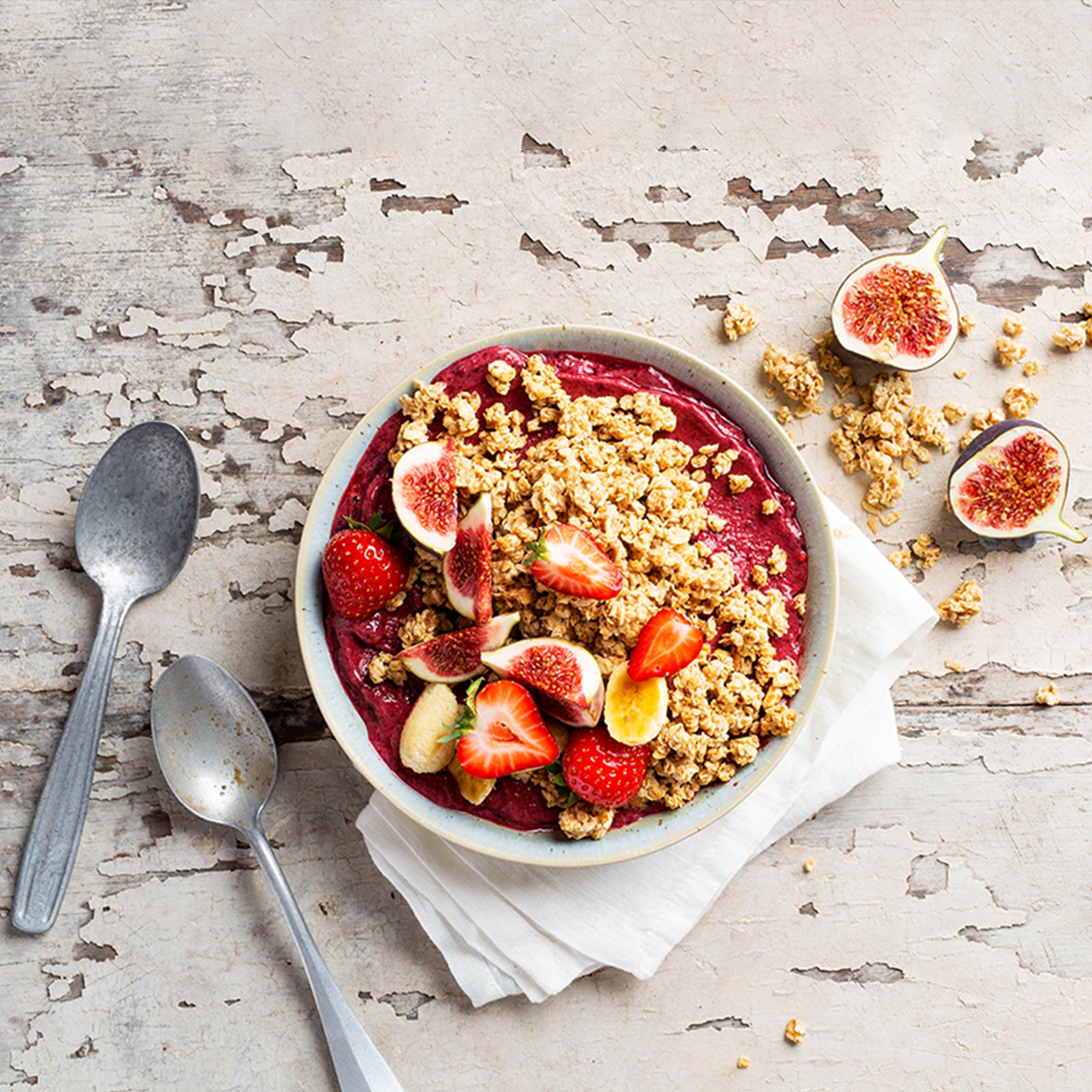 Ein Blick von oben auf eine wundervoll angerichtete Müsli-Bowl mit Beeren-Mousse und Krunchy Low Sugar Crazy Nuts von Bio-Knuspermüsli-Hersteller Barnhouse. Für ein optisches Highlight sorgt das Topping aus frischen Erdbeeren, Feigen und Bananen. Außerdem liegen noch zwei silberne Löffel, zwei Feigen-Hälfte und Müsli-Cluster auf dem Holztisch.