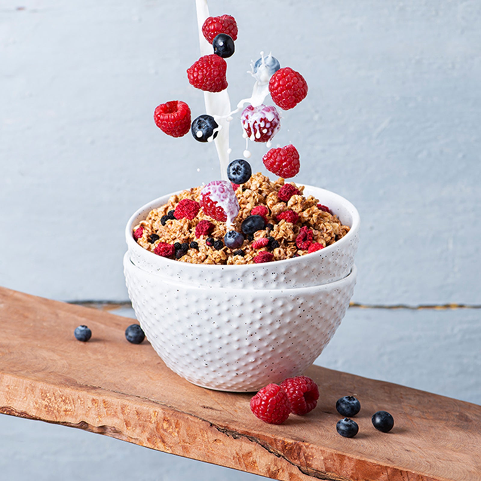 Blick von vorne auf zwei ineinander gestapelte, weiße Müslischalen, die auf einem leicht schrägen Holzbrett stehen. Daneben liegen Himbeeren und Heidelbeeren. Die obere Schüssel ist mit Krunchy Low Sugar Very Berry von Bio-Knuspermüsli-Hersteller Barnhouse befüllt, während von oben ein Milchstrahl sowie Himbeeren und Heidelbeeren ins Müsli fallen.