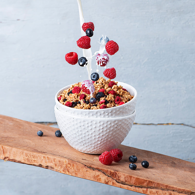 Blick von vorne auf zwei ineinander gestapelte, weiße Müslischalen, die auf einem leicht schrägen Holzbrett stehen. Daneben liegen Himbeeren und Heidelbeeren. Die obere Schüssel ist mit Krunchy Low Sugar Very Berry von Bio-Knuspermüsli-Hersteller Barnhouse befüllt, während von oben ein Milchstrahl sowie Himbeeren und Heidelbeeren ins Müsli fallen.