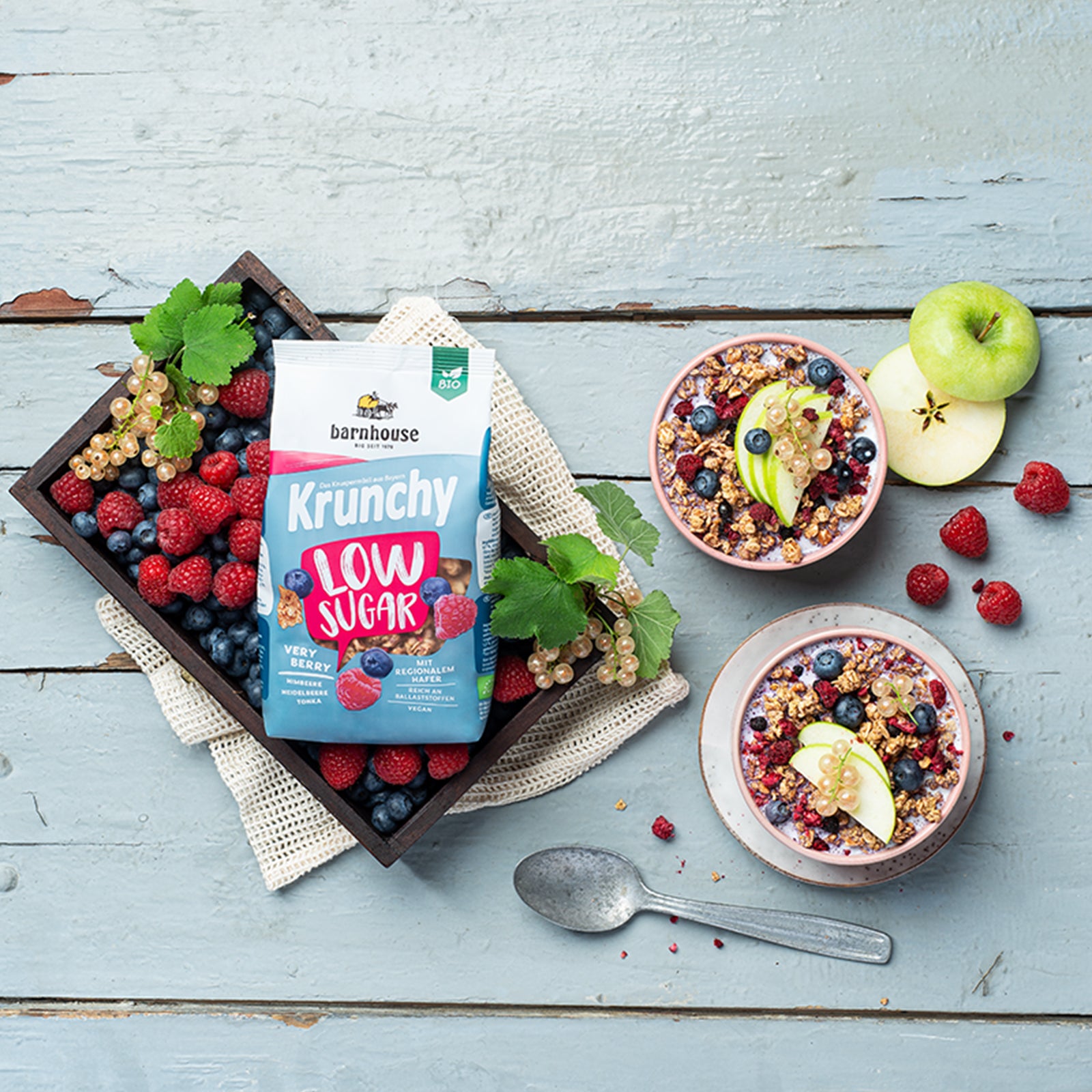Blick von oben auf einen rosa-weiß-blauen Beutel Krunchy Low Sugar Very Berry von Bio-Knuspermüsli-Hersteller Barnhouse, der in einer Kiste mit Himbeeren, Heidelbeeren und weißen Johannisbeeren auf einer Decke liegt. Daneben stehen 2 rosa Schüsseln (eine mit Unterteller) mit Milch & diesem zuckerarmen Müsli - jeweils mit Himbeeren, Heidelbeeren, weißen Johannisbeeren und grünen Apfelscheiben dekoriert. Auf dem bläulich gestrichenen Holztisch liegen ein silberner Löffel, ein geschnittener Apfel & Himbeeren.