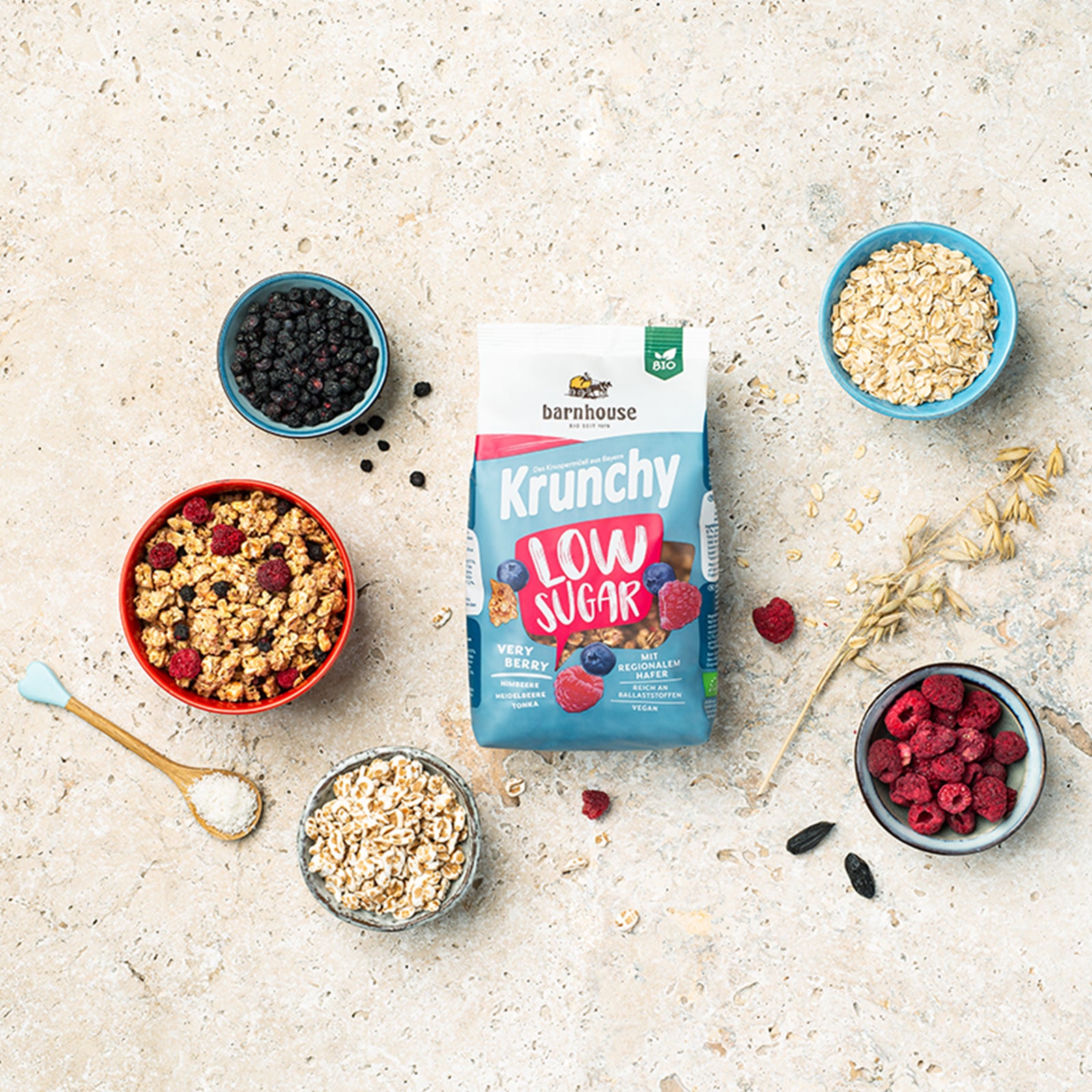 Blick von oben auf einen rosa-weiß-blauen Beutel Krunchy Low Sugar Very Berry von Bio-Knuspermüsli-Hersteller Barnhouse auf einer beigen Marmorplatte. Außenrum stehen vier verschiedenfarbige Schalen mit den Zutaten Heidelbeeren, Himbeeren, gepuffter Dinkel & Haferflocken sowie ein Holzlöffel mit Kokosraspeln. Eine weitere Schale ist mit dem beerigen, zuckerarmen Knuspermüsli befüllt. Ein Haferhalm liegt zwischen den Zutaten, die außerhalb der Schüsseln auf dem Tisch verteilt sind.
