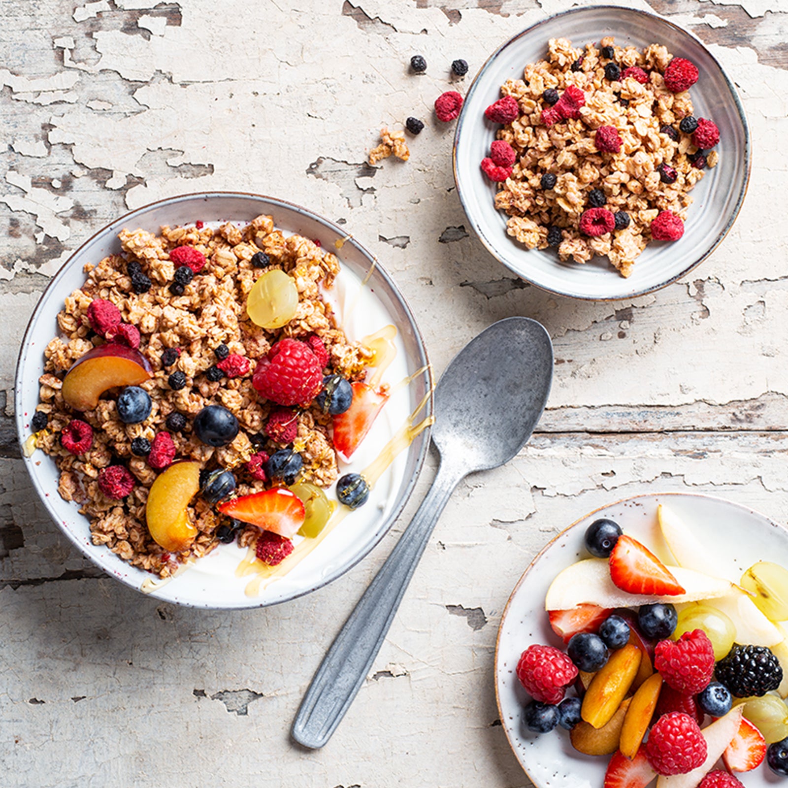 Etwas abgenutzter, grau angestrichener Holztisch, auf dem ein silberner Löffel liegt. Links steht eine blaue Müslischüssel mit Milch und Krunchy Low Sugar Very Berry von Bio-Knuspermüsli-Hersteller Barnhouse. Das Topping ist eine farblich abgestimmte Kombination aus Himbeeren, Heidelbeeren, Pfirsich und Weintrauben. Oben rechts steht eine Schale rein mit dem zuckerarmen Müsli. Unten rechts ist ein Teller mit Apfelscheiben, Erdbeeren, Himbeeren, Heidelbeeren, Pfirsich, Brombeeren und Weintrauben angerichtet.